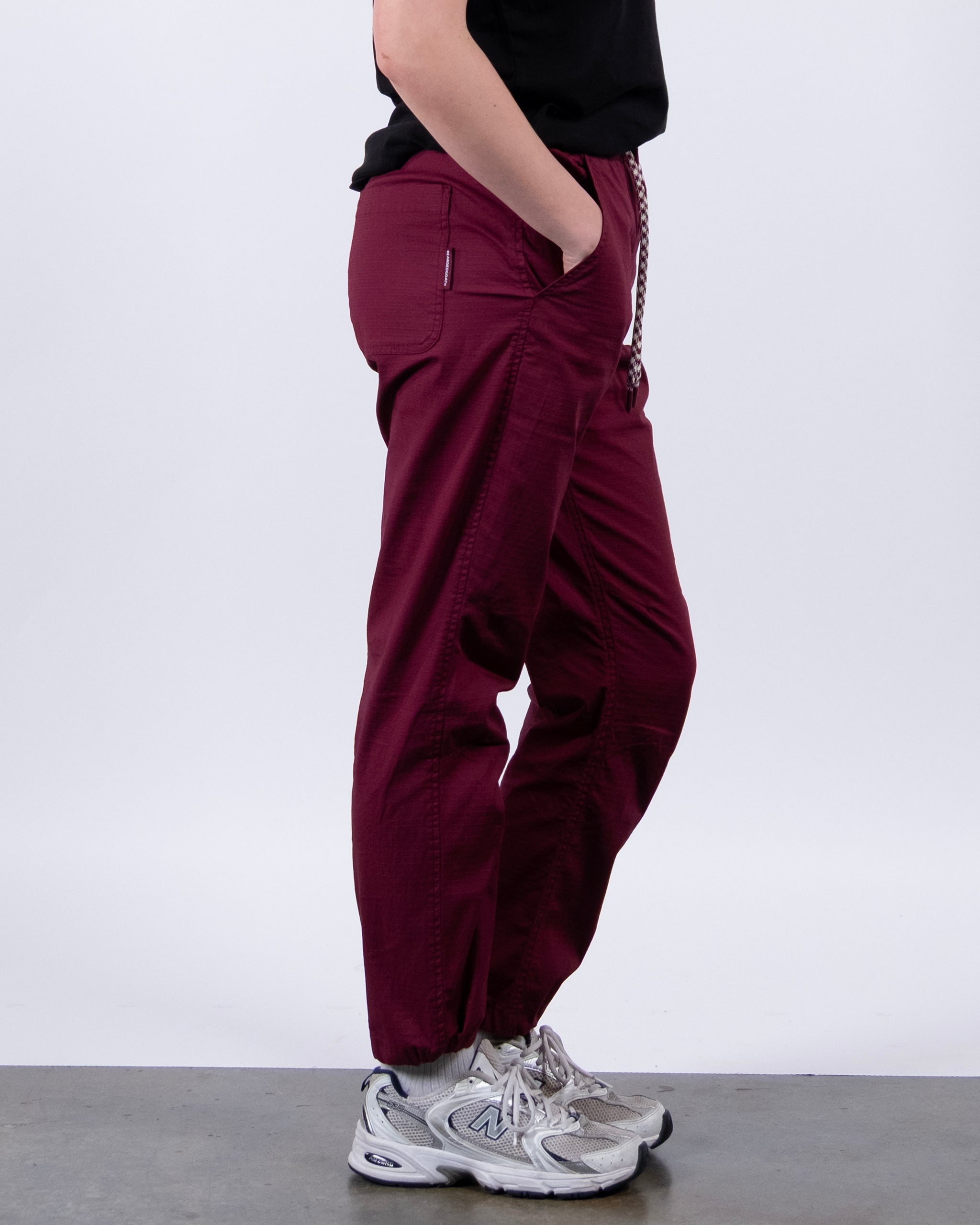 A person stands sideways against a white background wearing BEARDEDGOAT Women's Canopy™ Pant in maroon, made from stretch cotton ripstop, paired with a black short-sleeve shirt and gray sneakers. Their hand rests in the pant pocket.