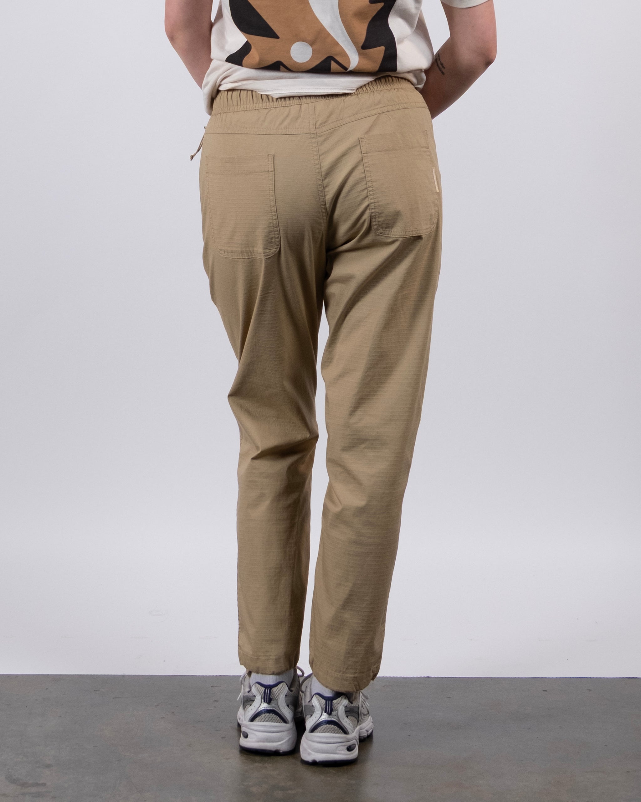 A person wears a graphic tee, BEARDEDGOAT Women's Canopy™ Pant with adjustable ankle drawcords, and white sneakers, standing facing away from the camera against a plain white background.