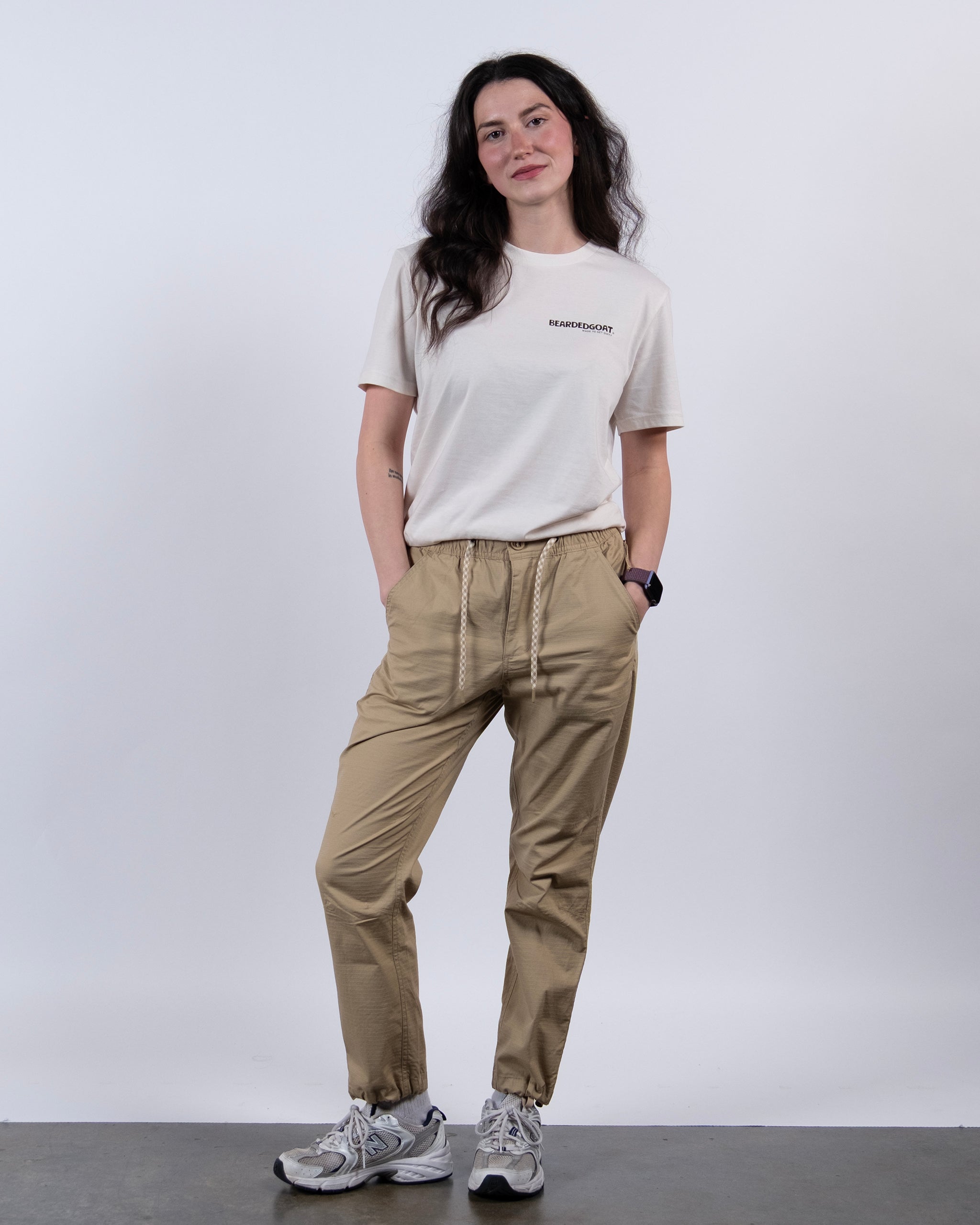 A woman with long dark hair stands against a white background, wearing a beige T-shirt, BEARDEDGOAT Women's Canopy™ Pant in stretch cotton ripstop with adjustable ankle drawcords, white sneakers, and a smartwatch, hands in pockets.