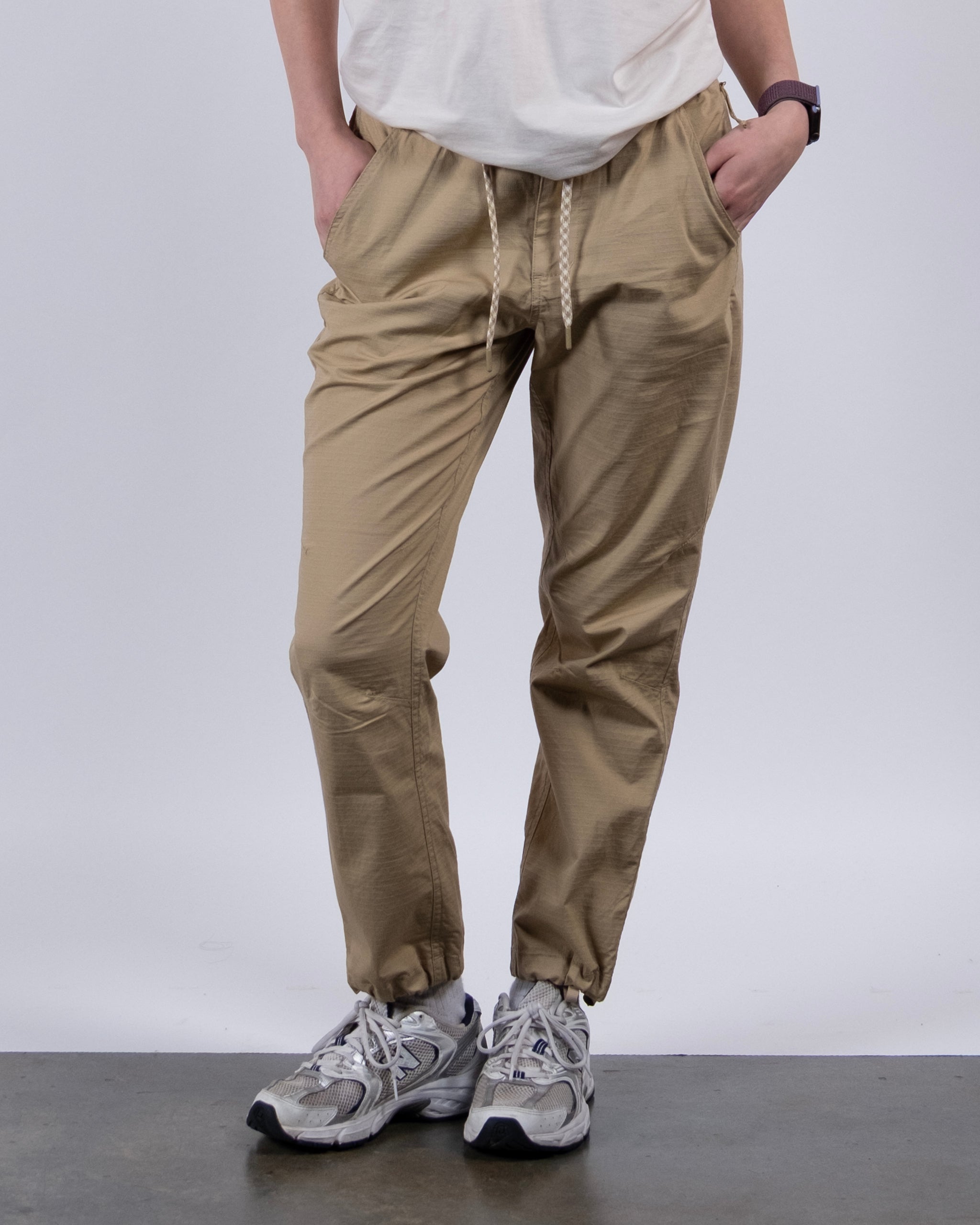 A person wears BEARDEDGOAT Women's Canopy™ Pants featuring adjustable ankle drawcords, paired with a white t-shirt, gray sneakers, and a watch, standing hands-in-pockets against a plain background.