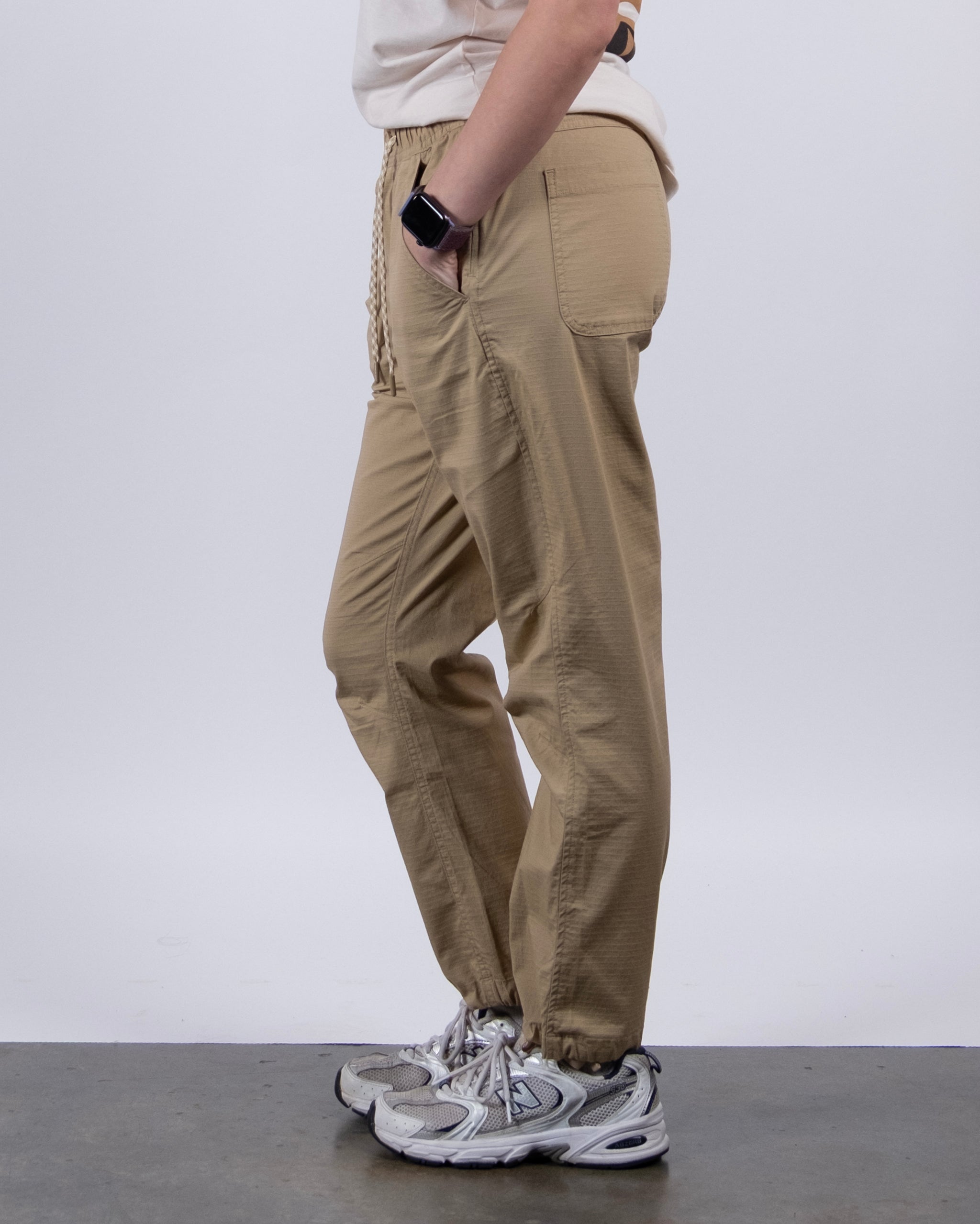 A person wearing BEARDEDGOAT Women's Canopy™ Pant in tan with adjustable ankle drawcords, a white T-shirt, smartwatch, and gray sneakers stands sideways on a gray floor against a plain white background.