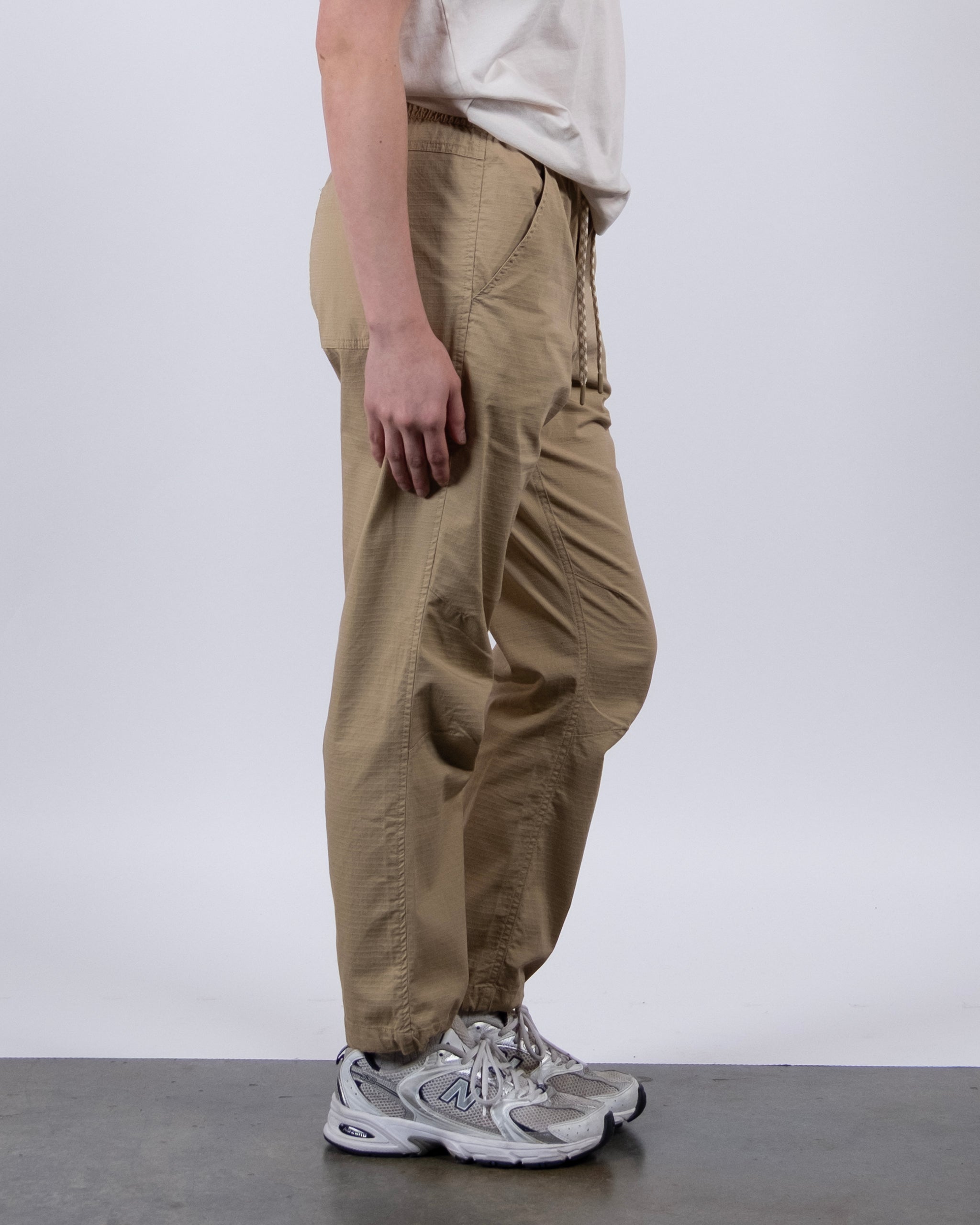 A person stands sideways against a light background wearing BEARDEDGOAT Women's Canopy™ Pant in tan, featuring adjustable ankle drawcords, paired with a white t-shirt and gray athletic shoes.