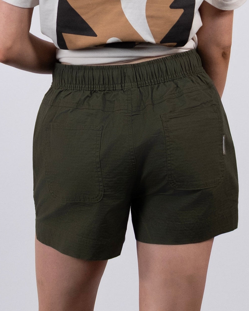 A person stands facing away, wearing BEARDEDGOAT Women's Canopy Short in olive green, a patterned t-shirt, white socks, and white sneakers against a plain light background.
