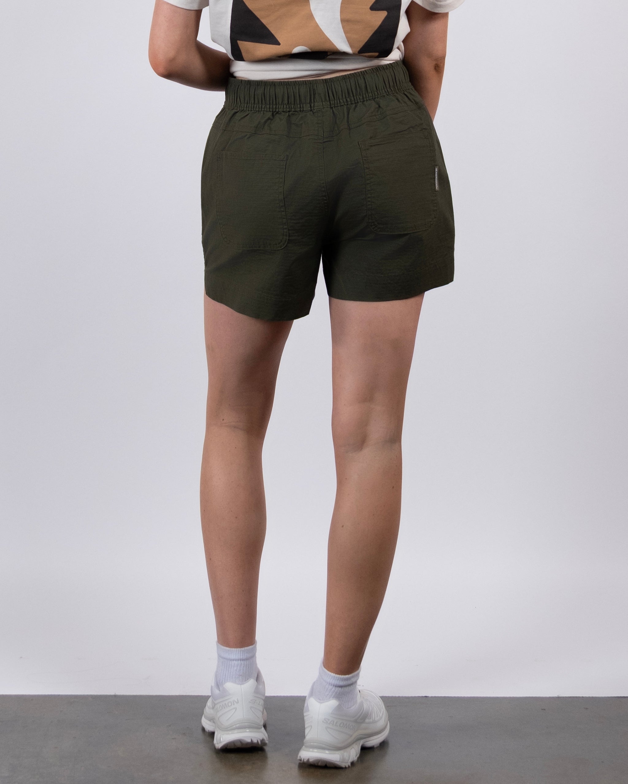 A person stands facing away, wearing BEARDEDGOAT Women's Canopy Short in olive green, a patterned t-shirt, white socks, and white sneakers against a plain light background.