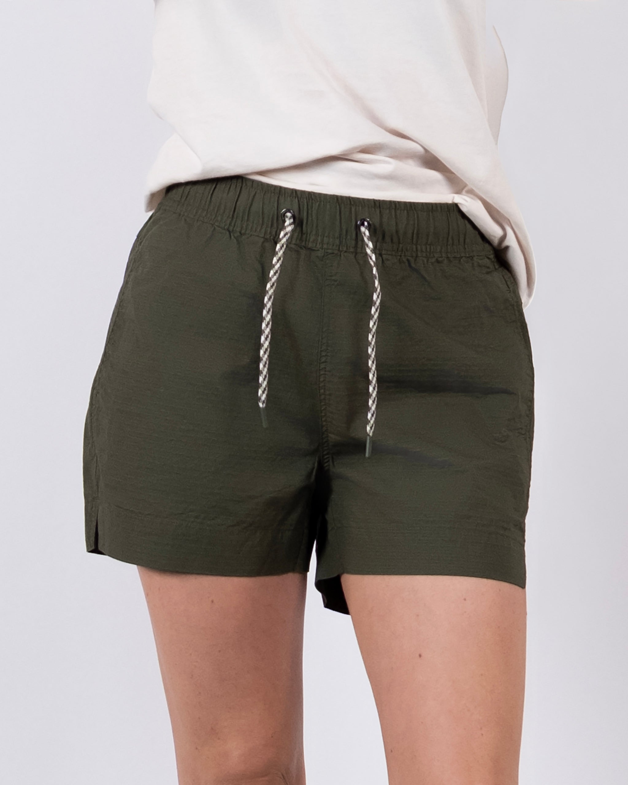 A person models BEARDEDGOAT's Women's Canopy Short in olive green with a drawstring, paired with a cream shirt. Only the lower torso and upper legs are visible against a plain white background.