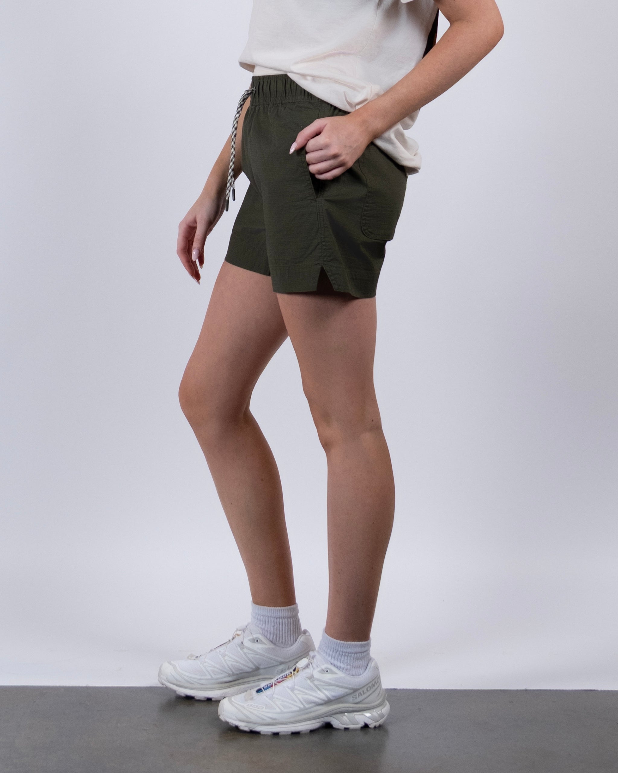 A person stands sideways in BEARDEDGOAT Women's Canopy Short, olive green stretch cotton ripstop with a drawstring, and a white t-shirt. White socks and sneakers complete the look against a plain light background.