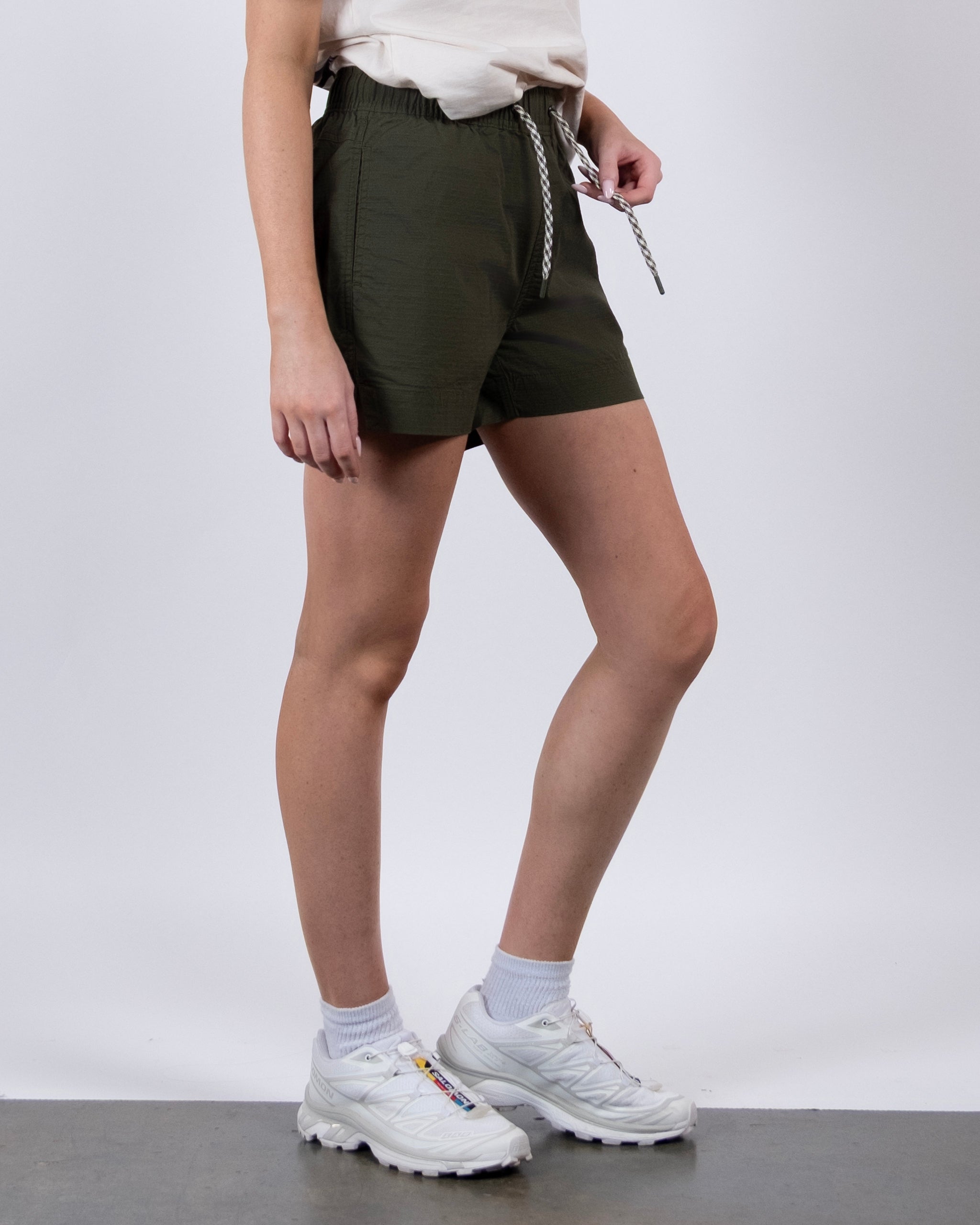 Only the lower half of a person is visible as they showcase BEARDEDGOAT Women's Canopy Shorts in olive green athletic fit stretch cotton ripstop, styled with a white t-shirt, socks, and sneakers against a plain background.