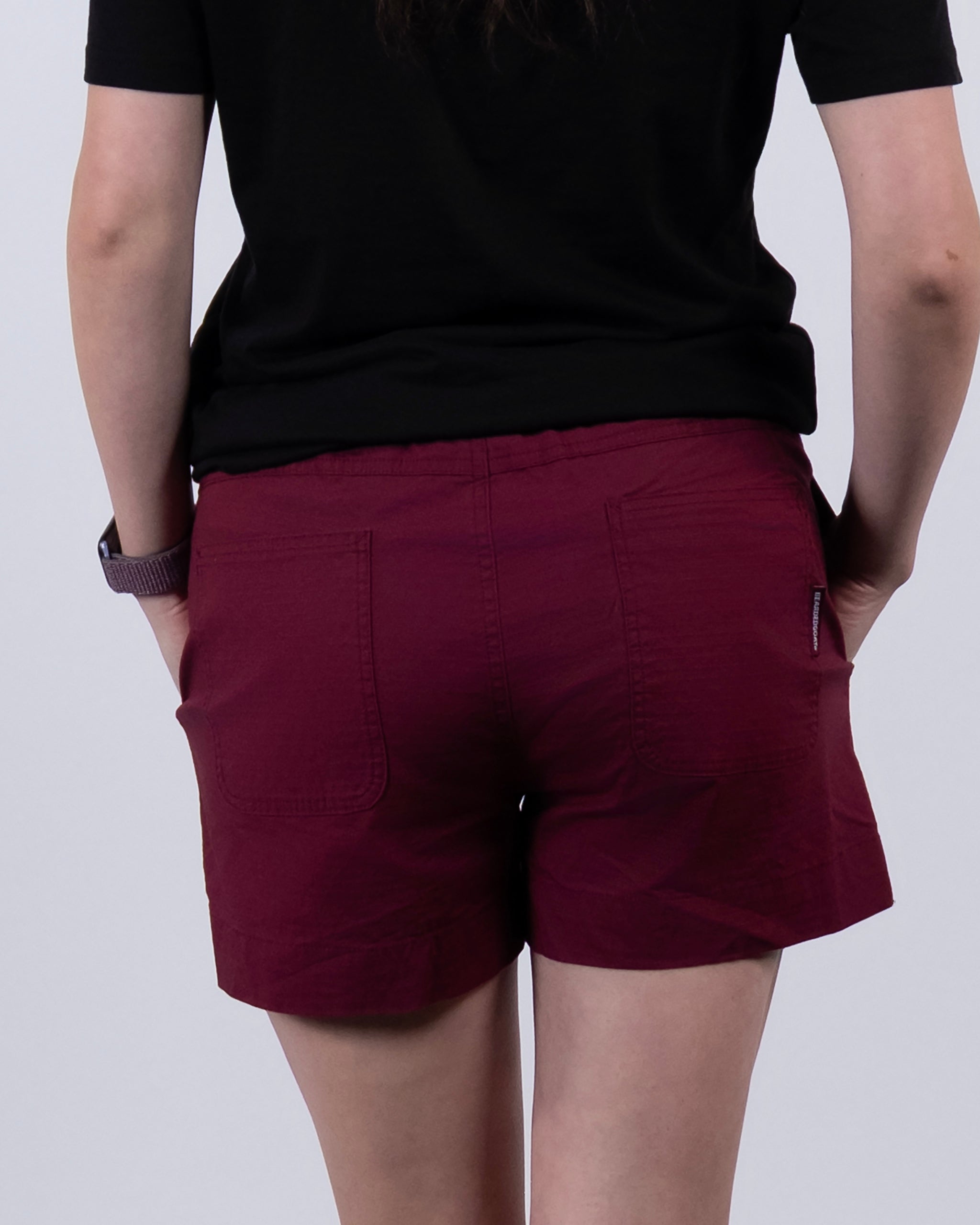 A person wearing an athletic fit black shirt and BEARDEDGOAT Women's Canopy Short in maroon stands with their back to the camera, hands in pockets, against a plain light background.