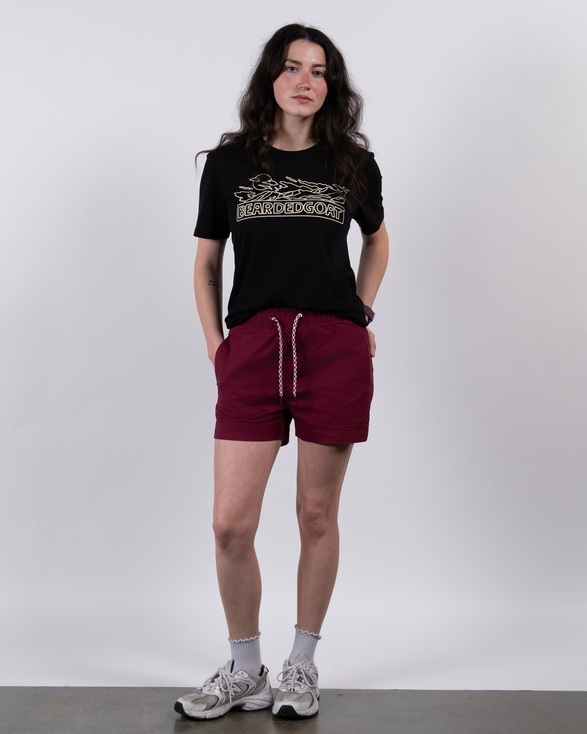 A woman with long dark hair stands against a white background, wearing BEARDEDGOAT Women's Canopy Shorts in maroon, a black graphic T-shirt, white socks, and gray sneakers, with her hands in her shorts pockets.