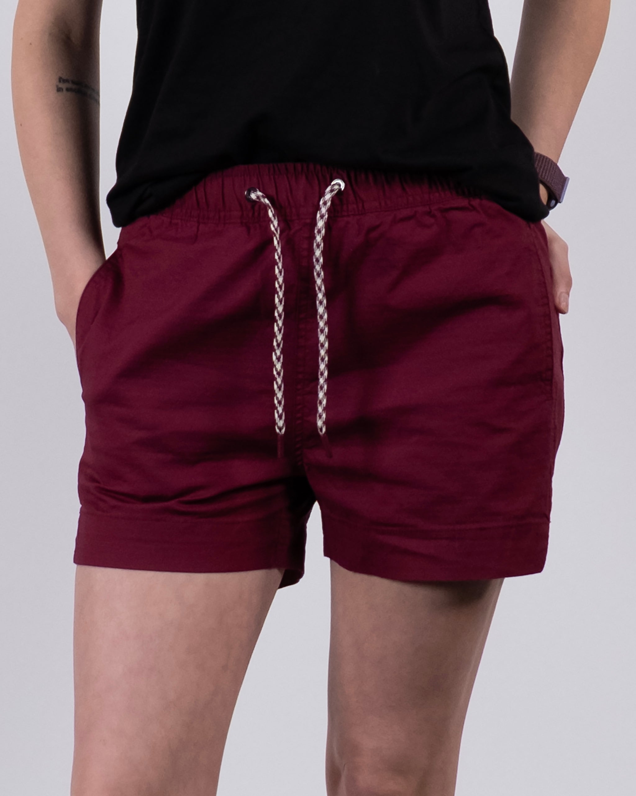 A person wearing BEARDEDGOAT Women's Canopy Short in burgundy, made from stretch cotton ripstop for breathable comfort with a white and black drawstring, paired with a black shirt and watch, stands hands in pockets.