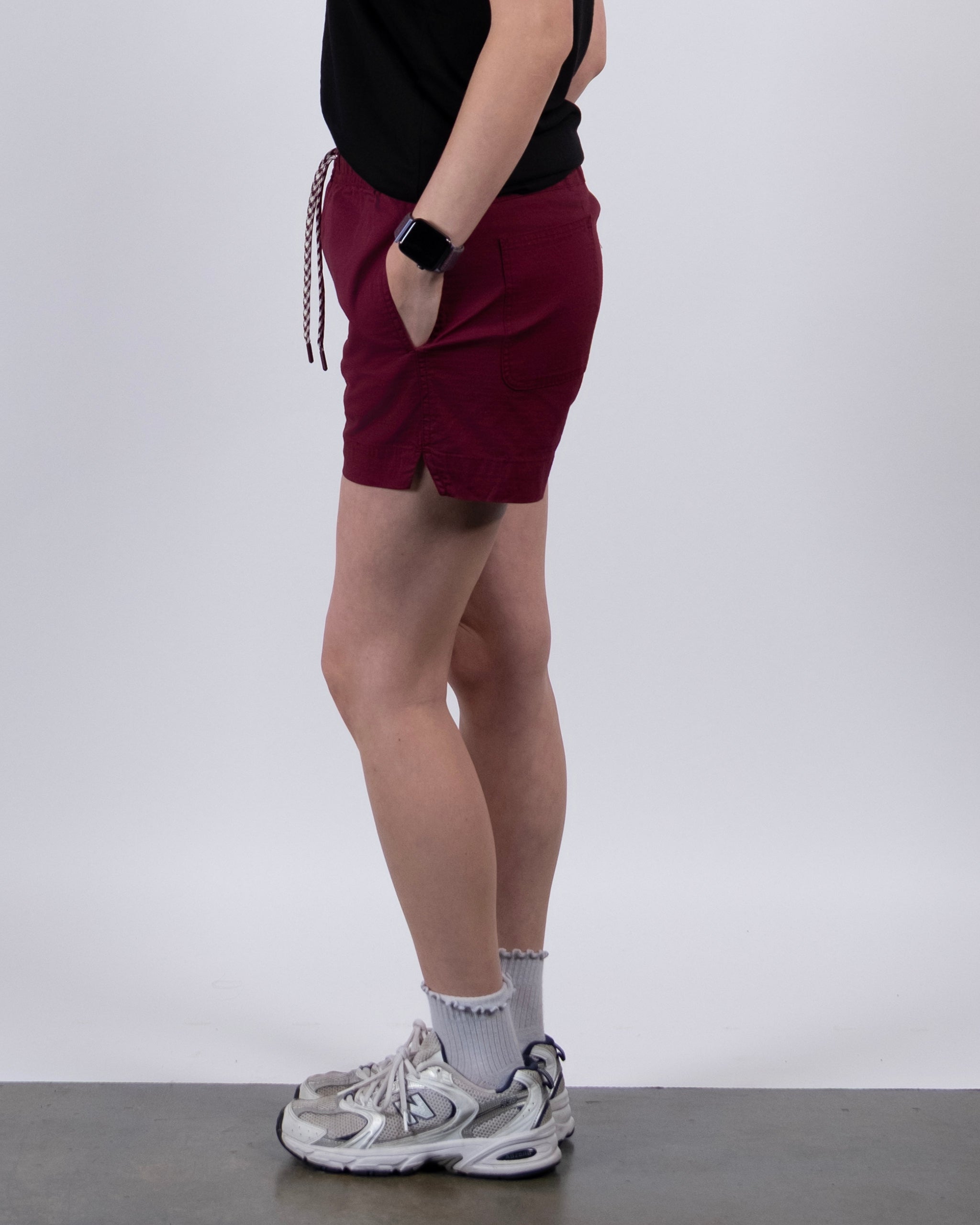A person standing sideways in a black shirt, BEARDEDGOAT Women's Canopy Short in maroon with pockets, white athletic socks, gray sneakers, and a smartwatch against a plain white background.
