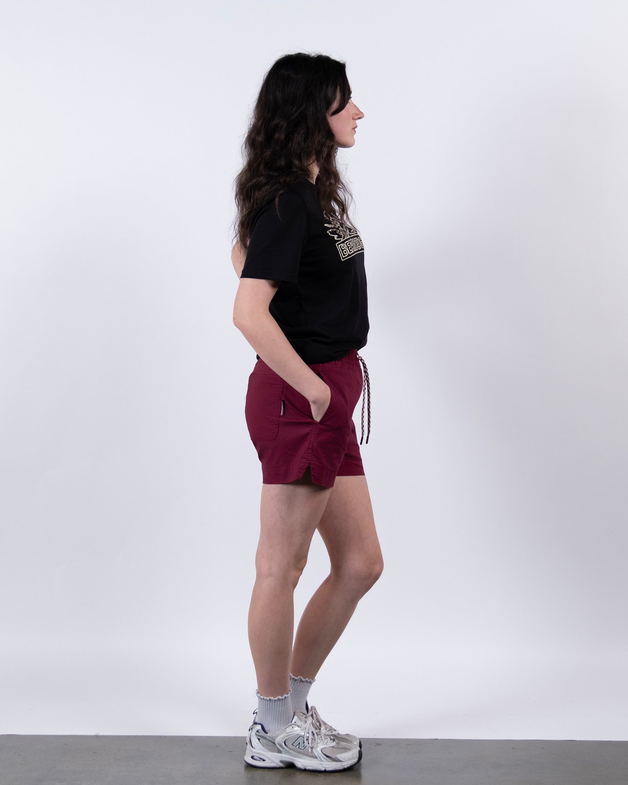 A woman with long, wavy dark hair stands in profile against a white background, hands in pockets, wearing BEARDEDGOAT Women's Canopy Short in maroon, a black t-shirt, white socks, and white sneakers.