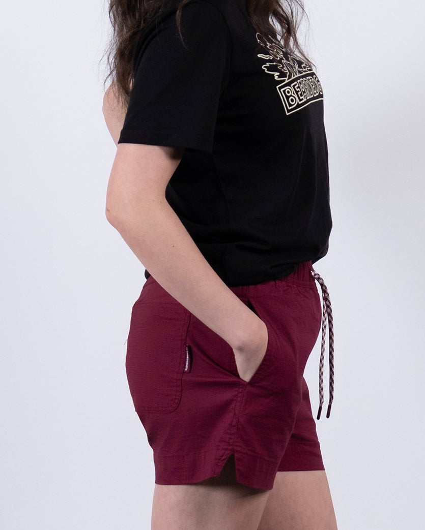 A woman with long, wavy dark hair stands in profile against a white background, hands in pockets, wearing BEARDEDGOAT Women's Canopy Short in maroon, a black t-shirt, white socks, and white sneakers.