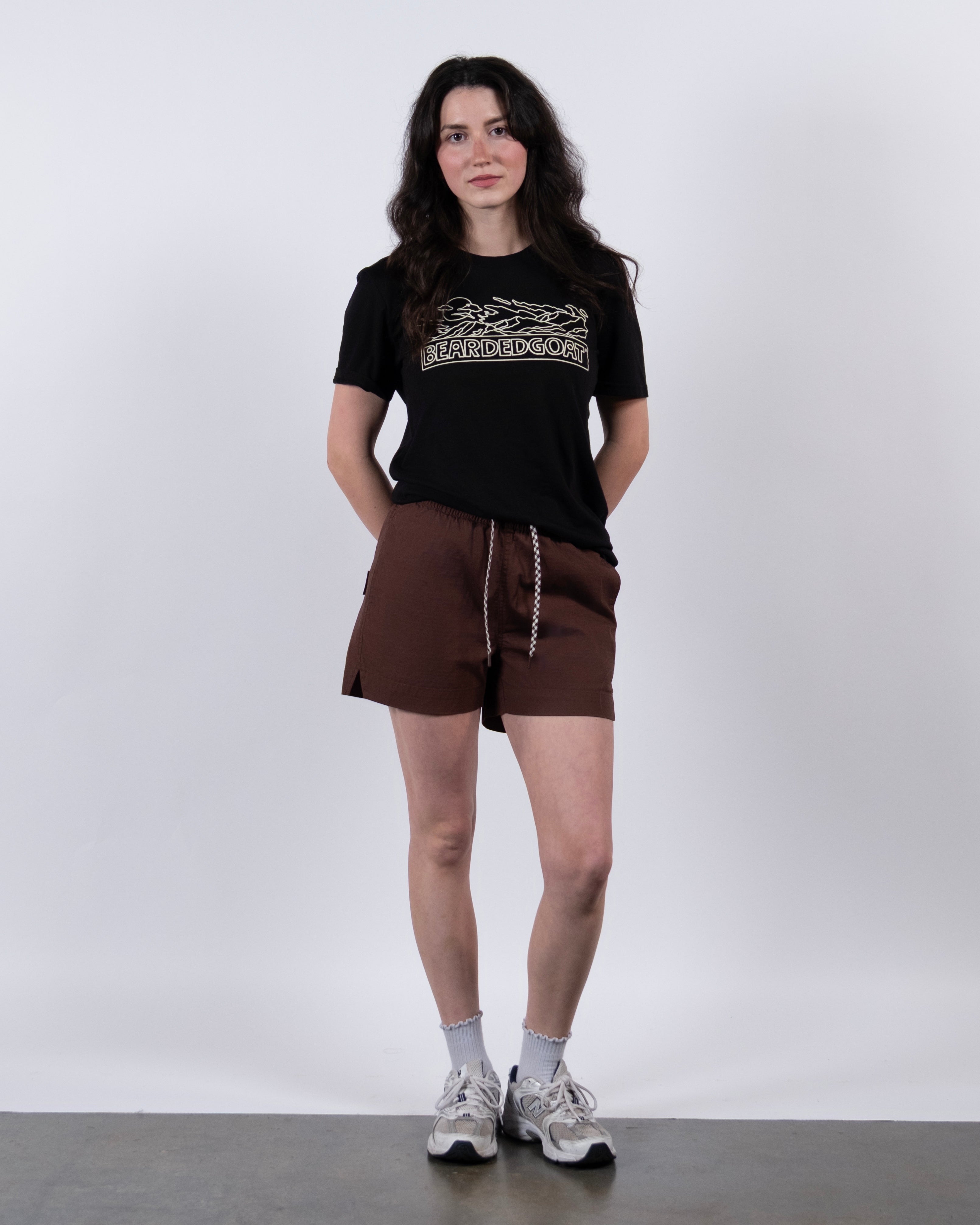 A person with long dark hair stands against a white background, wearing a black graphic tee, BEARDEDGOAT Women's Canopy Short in brown cotton ripstop with a drawstring waist, white socks, and white sneakers. Hands rest in their shorts pockets.