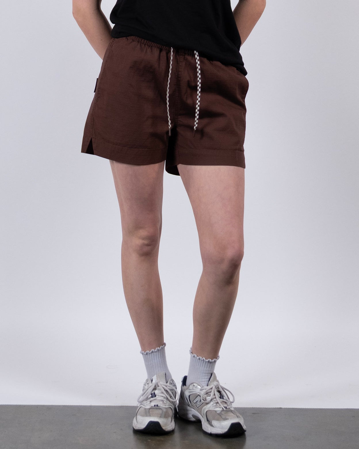 A person wears BEARDEDGOAT Women's Canopy Short in brown stretch cotton ripstop, paired with a black t-shirt, white socks, and white sneakers. The cropped photo shows the outfit from shoulders down against a light gray background.