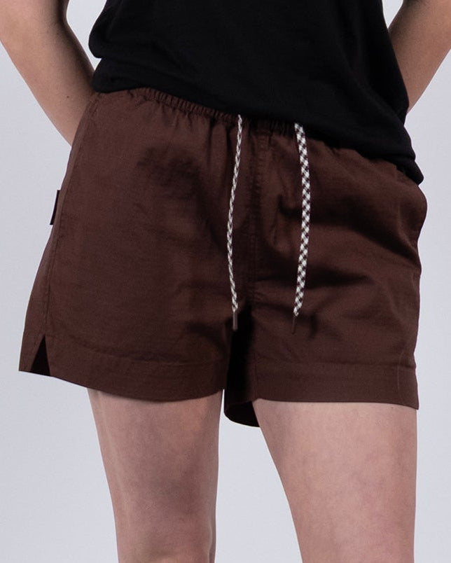 A person wears BEARDEDGOAT Women's Canopy Short in brown stretch cotton ripstop, paired with a black t-shirt, white socks, and white sneakers. The cropped photo shows the outfit from shoulders down against a light gray background.