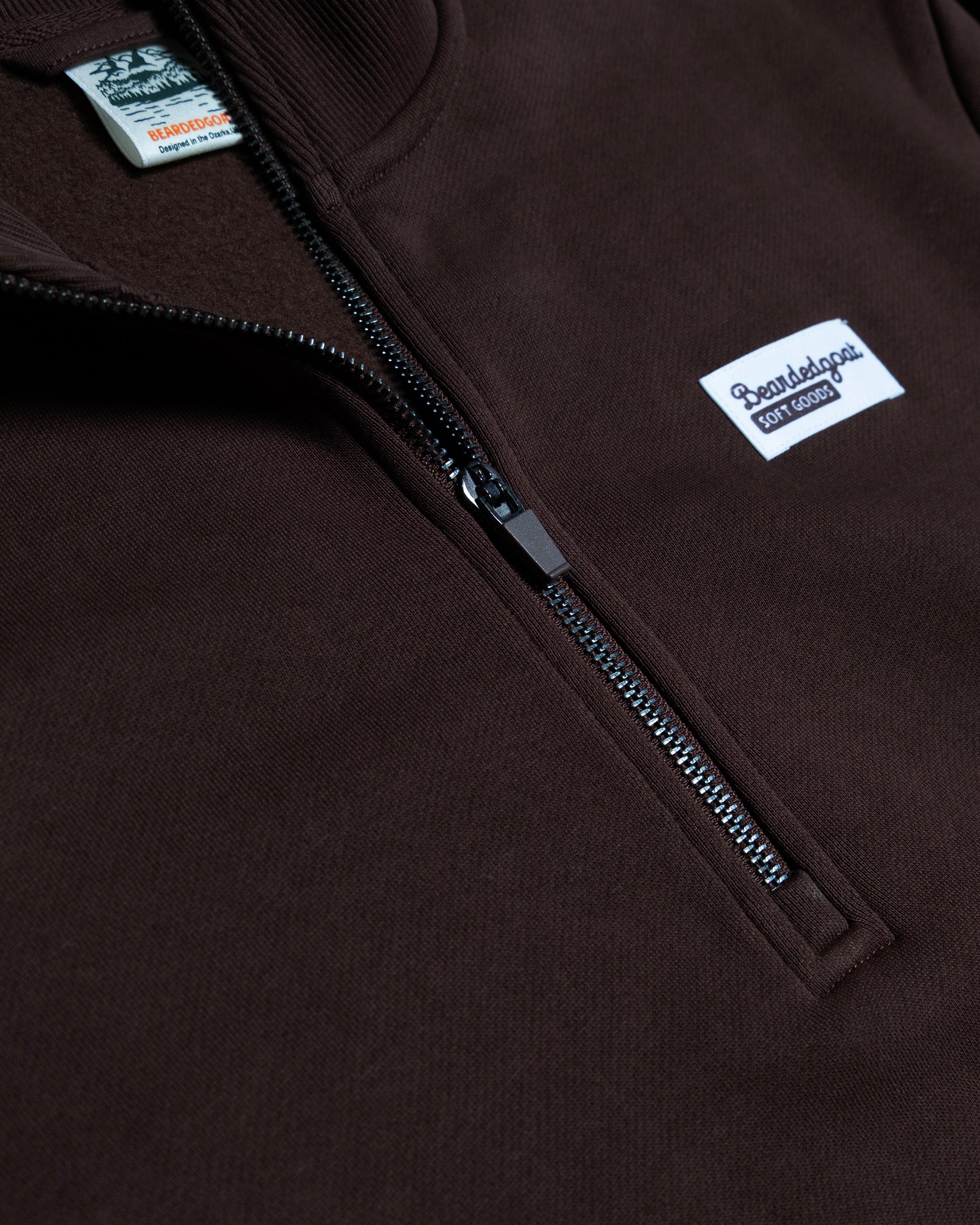 Close-up of the BEARDEDGOAT NEW — VOID™ Half Zip: a brown heavyweight fleece quarter-zip with a black zipper and a white Raised by Gods Soft Goods patch on the chest. Partially visible clothing tags appear near the collar.