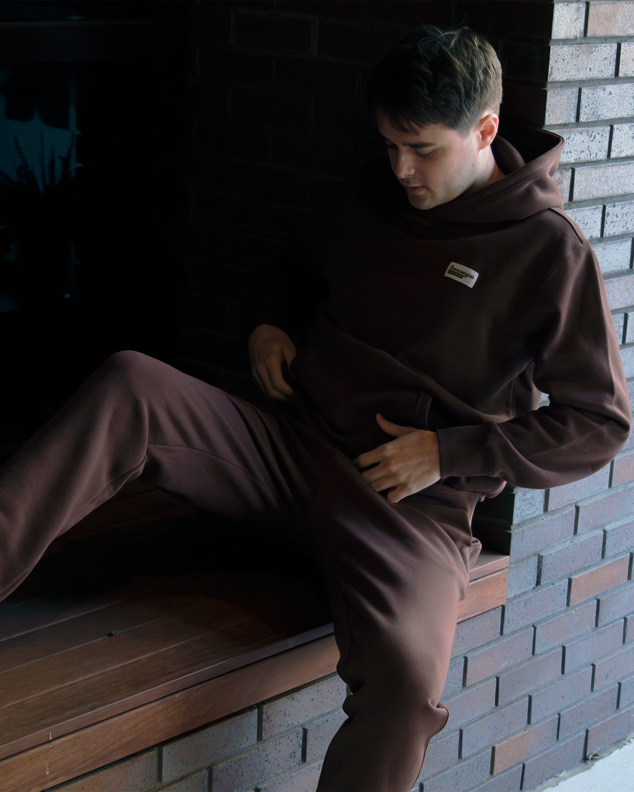 Seated on wooden steps by a brick wall, a person in the beardedgoat VOID™ Hoodie and matching pants looks down, hands in their lap. Soft light highlights the premium heavyweight cotton fleece of the apparel.