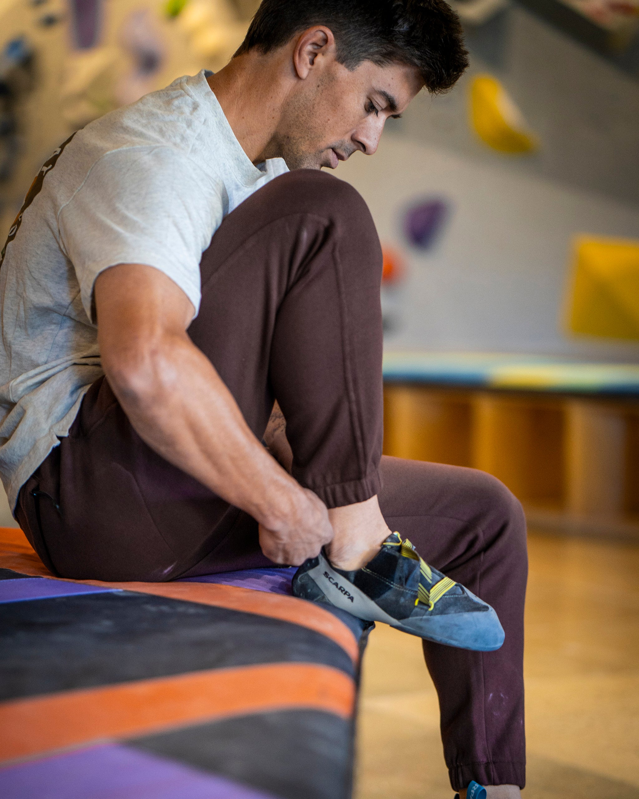 A man in a light t-shirt and beardedgoat VOID™ Sweatpant sits on a padded bench, putting on blue and gray climbing shoes. Blurred climbing holds and gym features appear in the background.