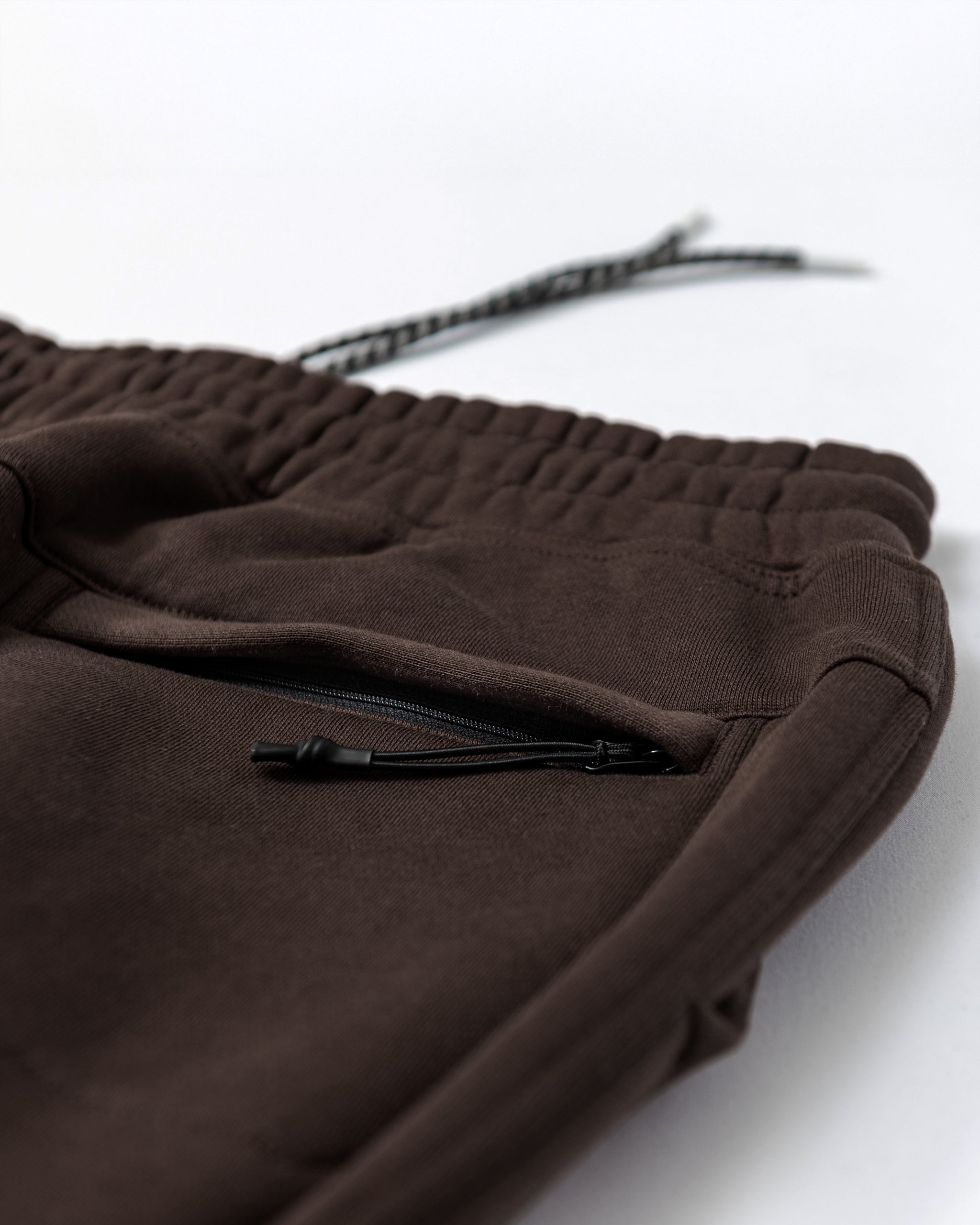 Close-up of durable beardedgoat VOID™ Sweatpant in brown, featuring an elastic waistband, drawstring, and a zippered pocket with black zipper pull, displayed on a white surface.