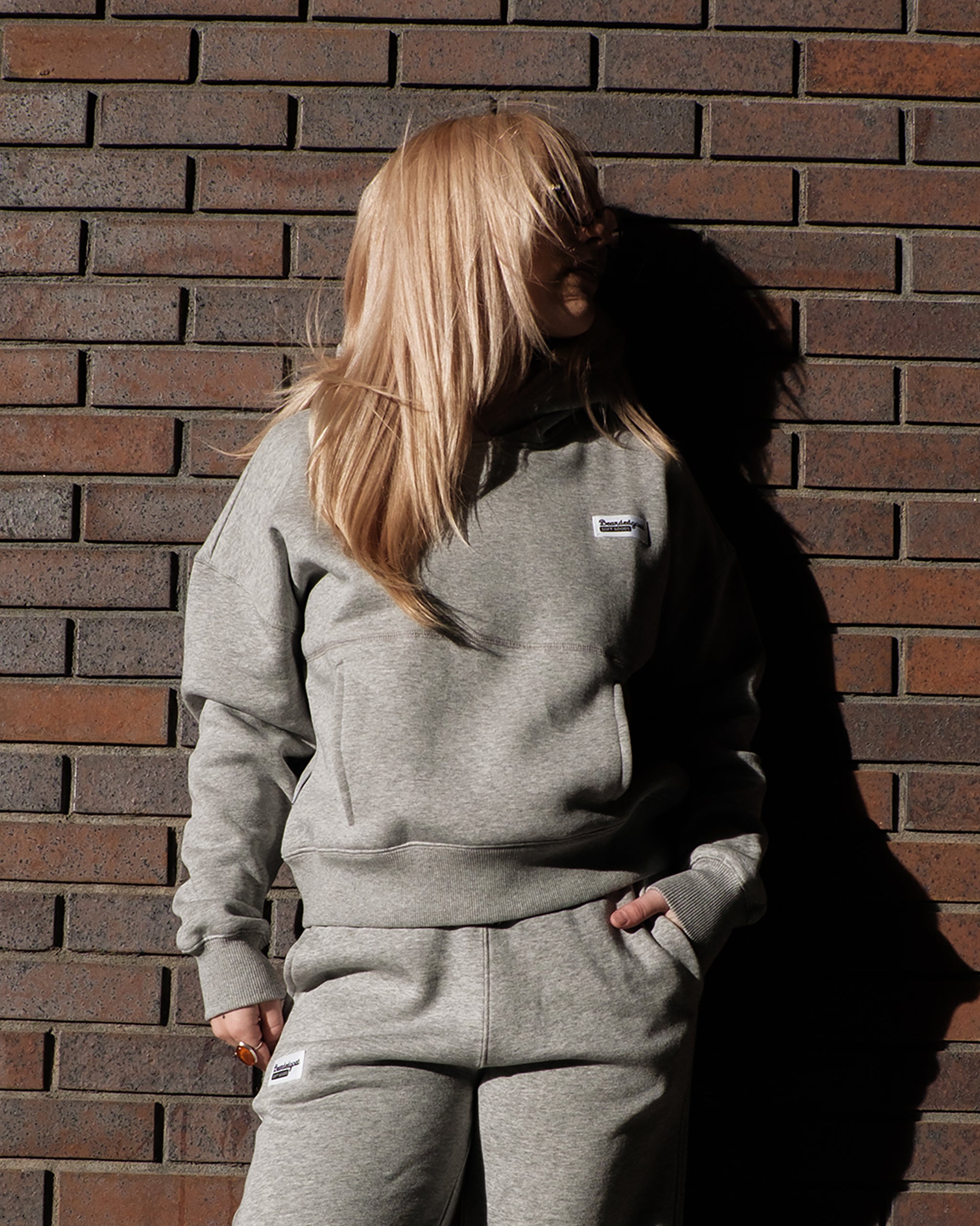 A woman with long blonde hair stands by a brown brick wall, wearing the beardedgoat Women's Overnight™ Hoodie in light gray, made from premium cotton fleece. Strong lighting casts dramatic shadows behind her.