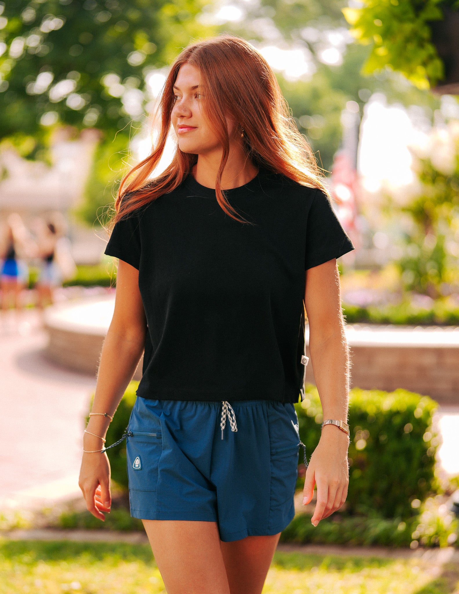 A young woman with long red hair wearing a black t-shirt and BEARDEDGOAT Summit Shorts, crafted from lightweight ripstop fabric, walks outdoors in a sunny park surrounded by greenery and blurred figures in the background.