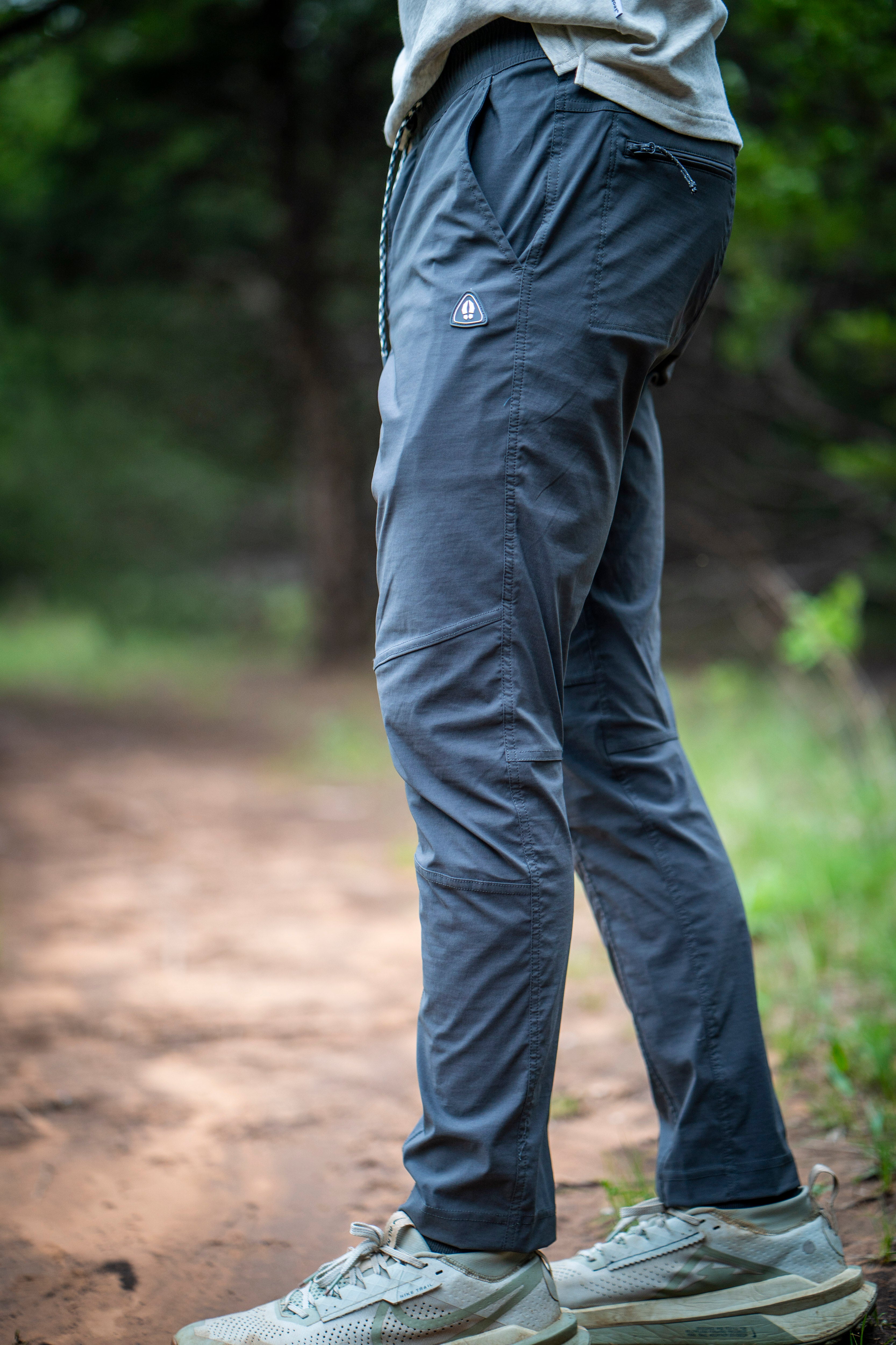 A person wearing BEARDEDGOAT Mens Rover Pant, made with stretch water-resistant fabric, and light trail shoes stands on a dirt path in a lush wooded area. Photo focuses on the pants and shoes from the waist down.