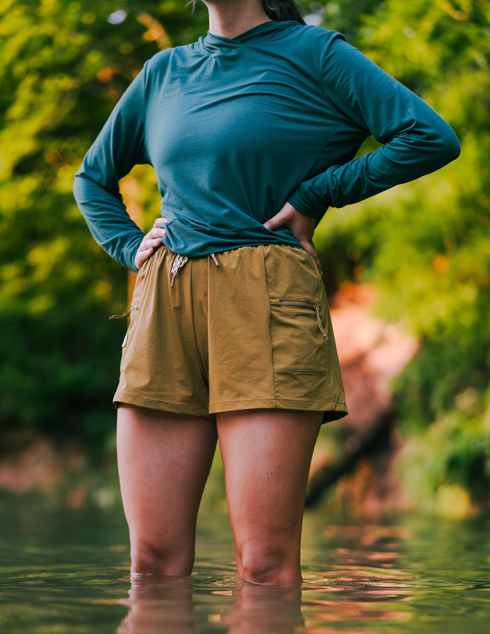 A person stands in shallow water with hands on hips, wearing a teal long-sleeve shirt and tan high-waisted adventure short made from ripstop fabric. Green leafy trees fill the blurred background.