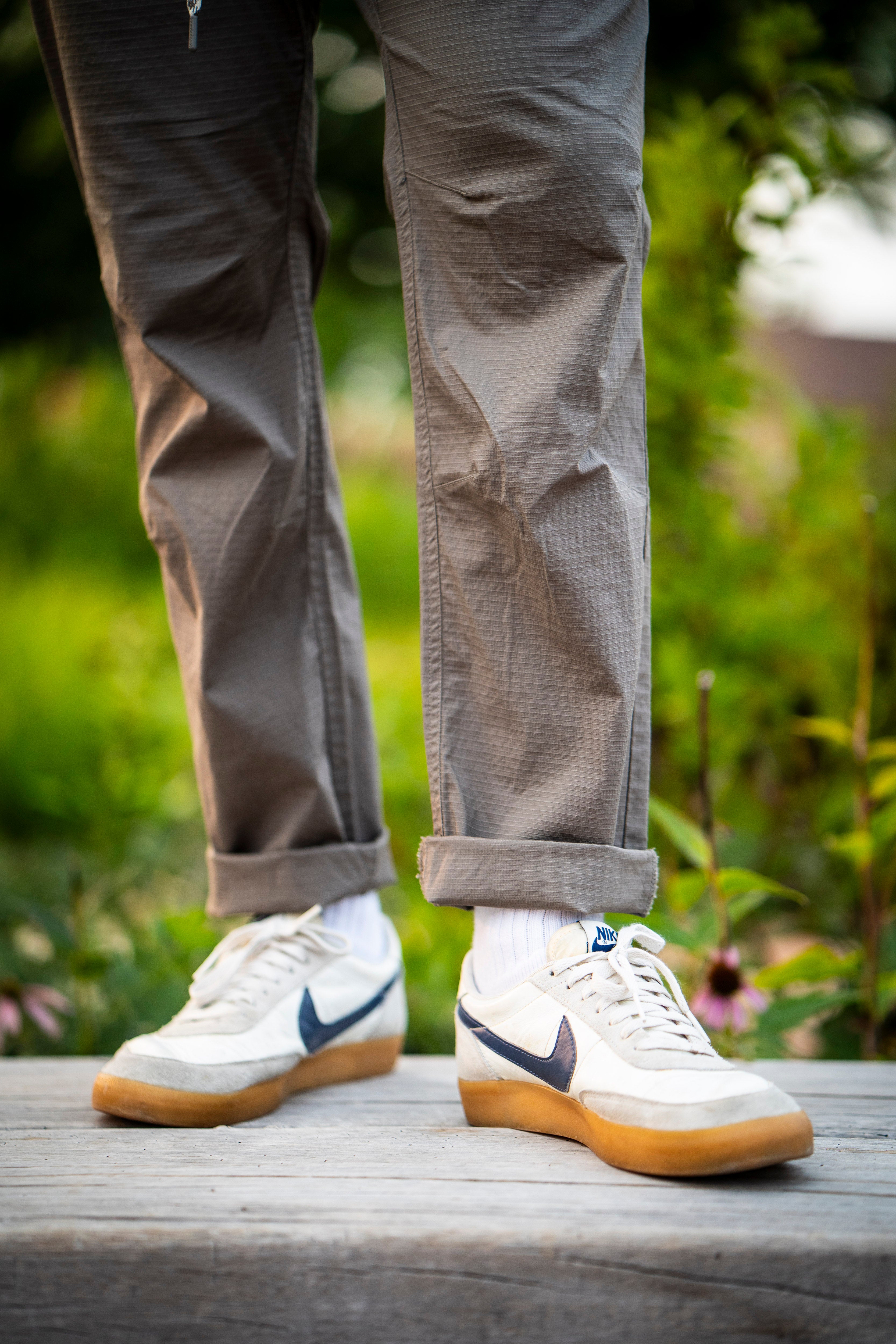 A person stands outdoors on wooden planks, wearing BEARDEDGOAT Men's Canopy Pant in light gray, cuffed at the ankle, with white Nike sneakers and white socks. Blurred greenery and purple flowers appear in the background.