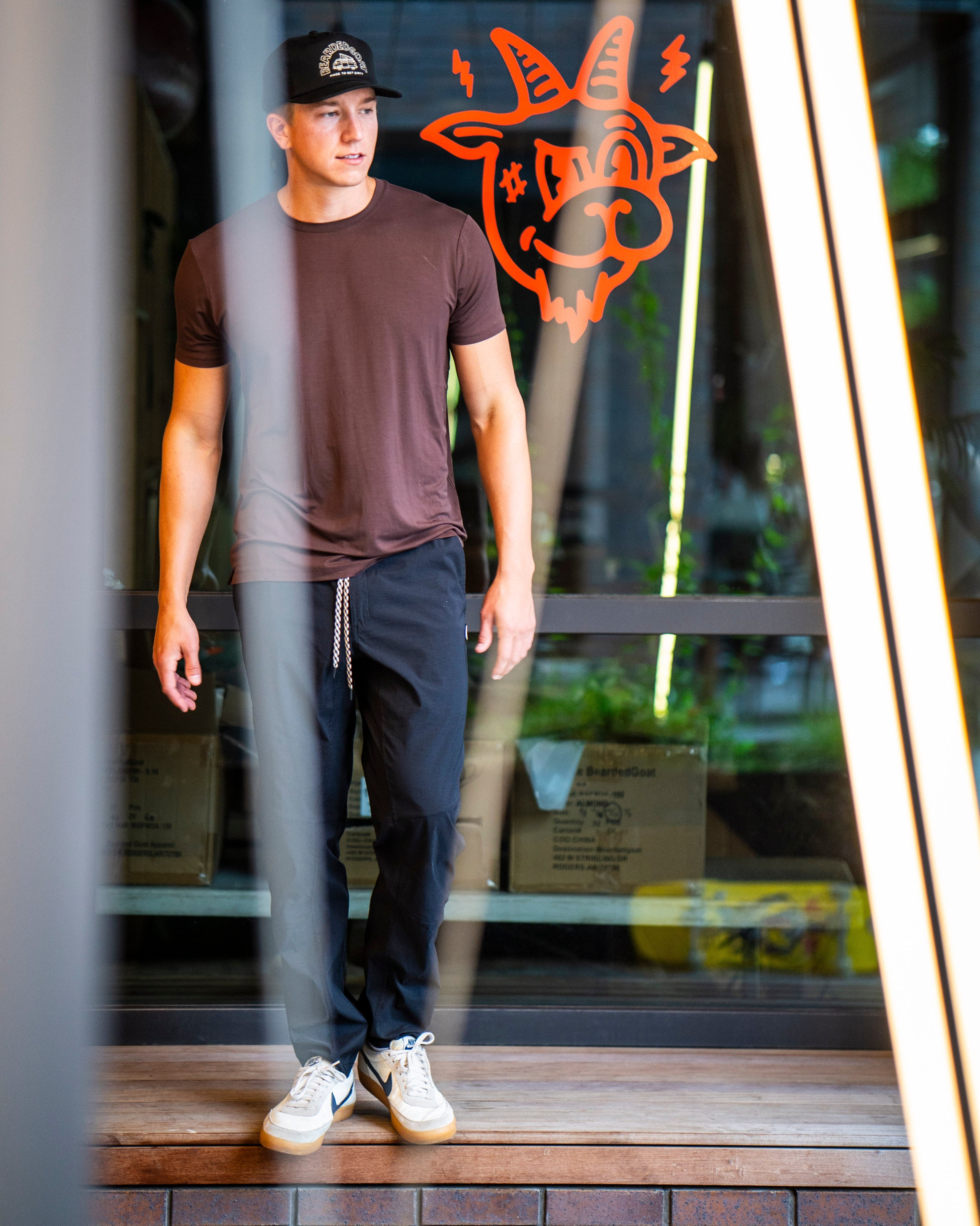 A man in a brown T-shirt, BEARDEDGOAT NEW-—ROVER™ Pant, and white sneakers stands near a window with an orange cartoon goat face decal. Cardboard boxes are visible behind the glass. Bright diagonal beams frame the image.