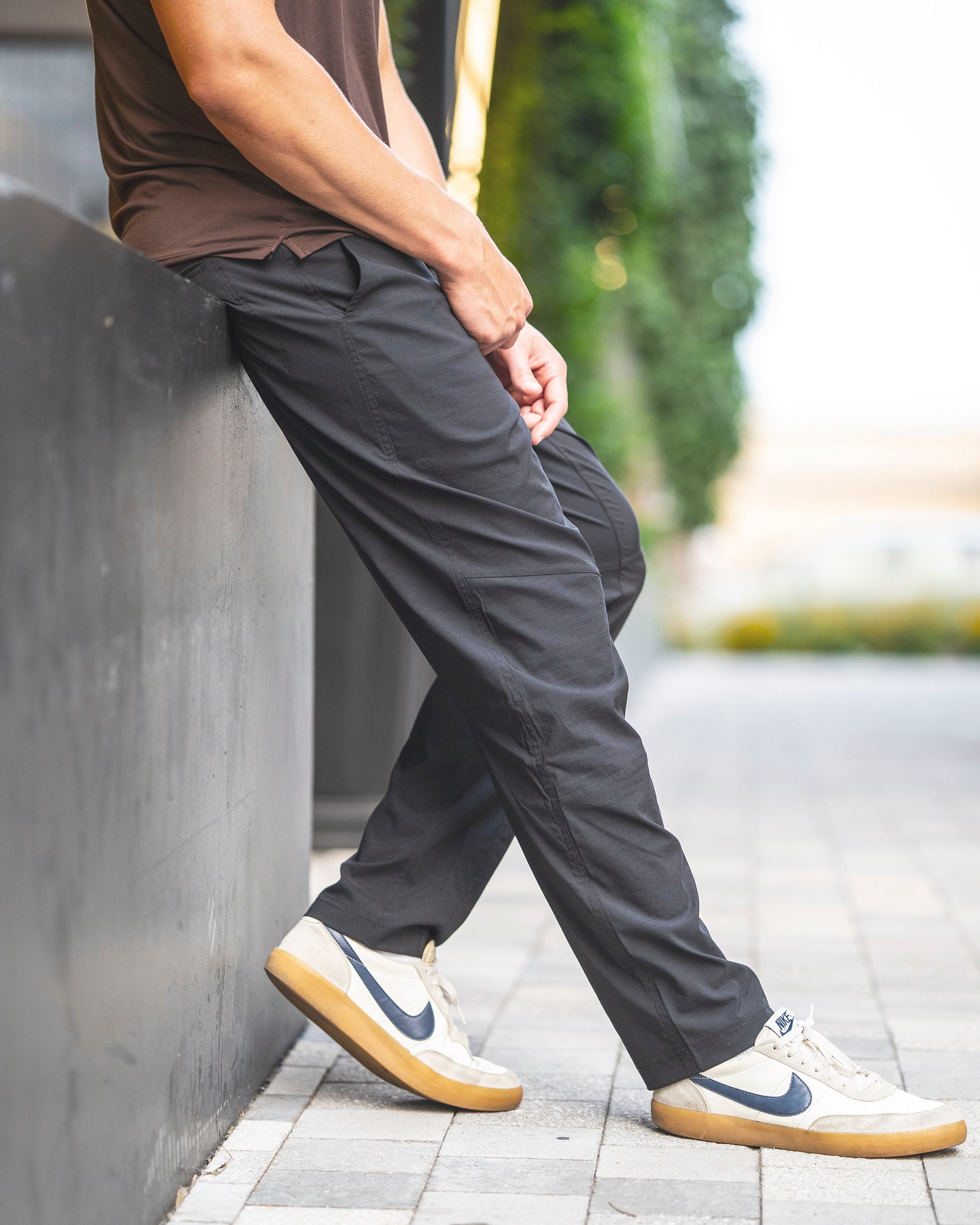A person in black BEARDEDGOAT NEW-—ROVER™ Pants, a brown shirt, and white Nike sneakers with black swooshes leans casually against a black wall on a paved walkway, greenery in the background.