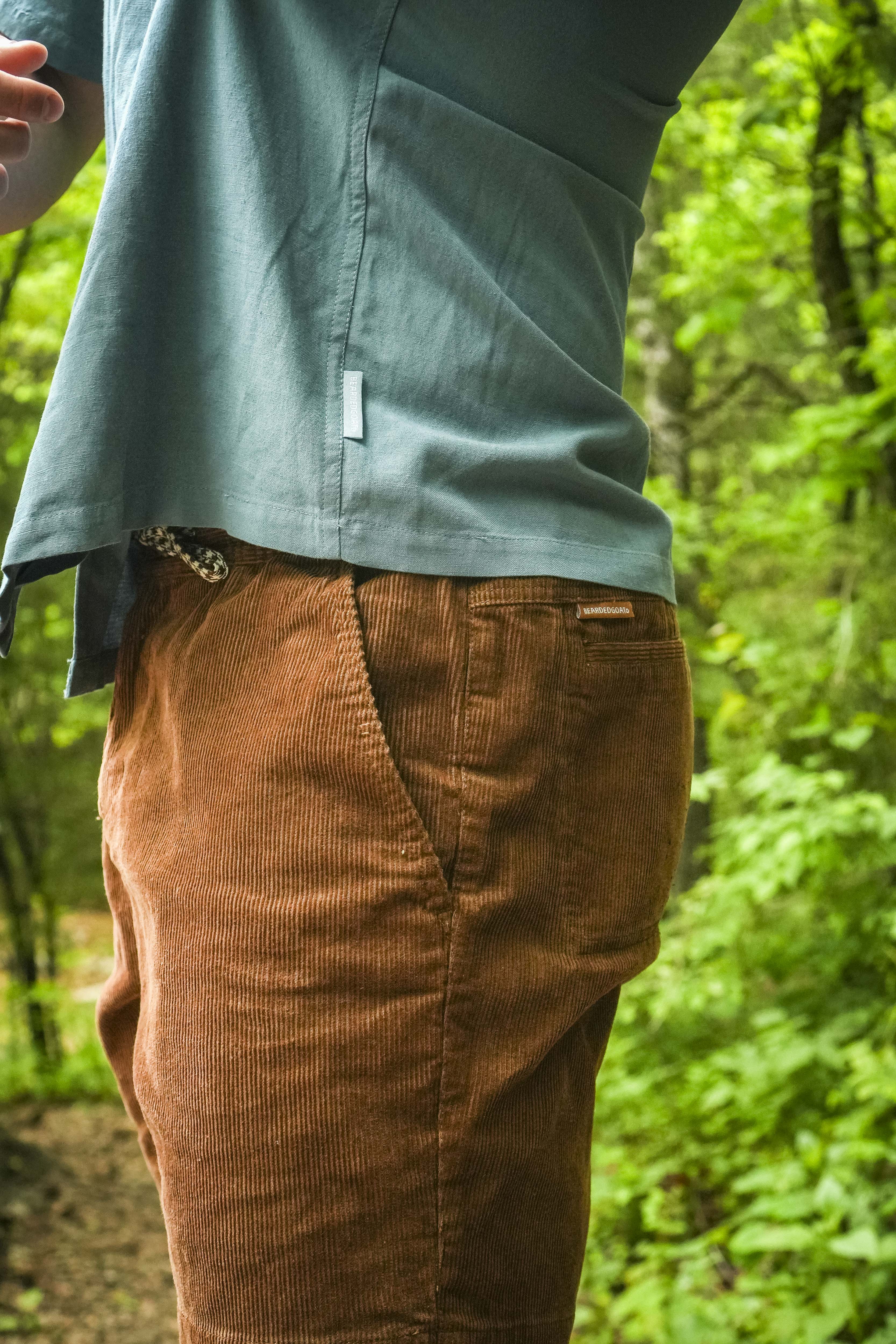 A person wearing BEARDEDGOAT Mens Cord Shorts stands on a forest trail in a blue shirt, surrounded by lush trees and greenery. The image is cropped to highlight the shorts and upper body.