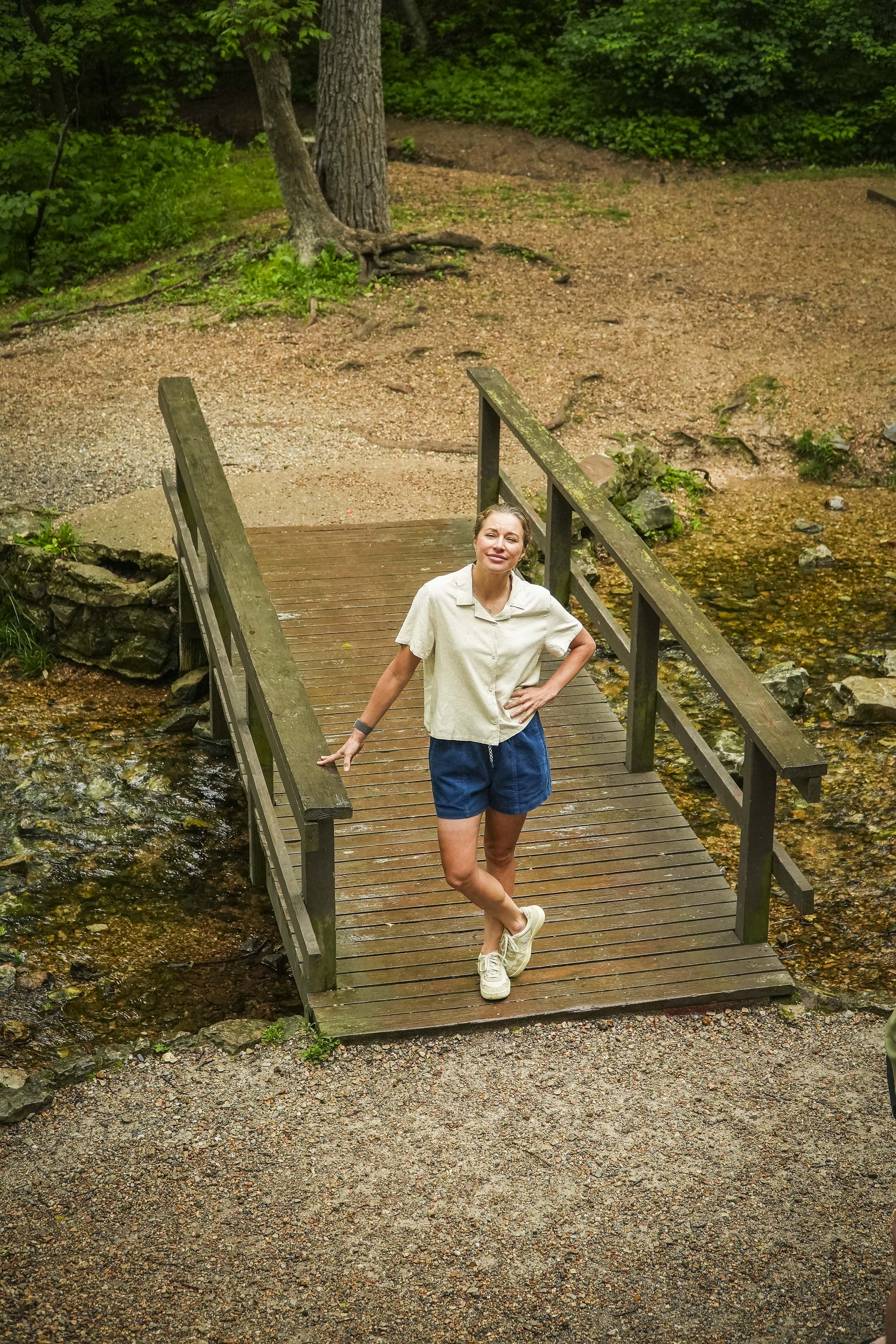 A woman wearing BEARDEDGOAT Womens Cord Shorts and a light shirt stands smiling on a wooden footbridge over a small stream, surrounded by greenery and gravel paths.