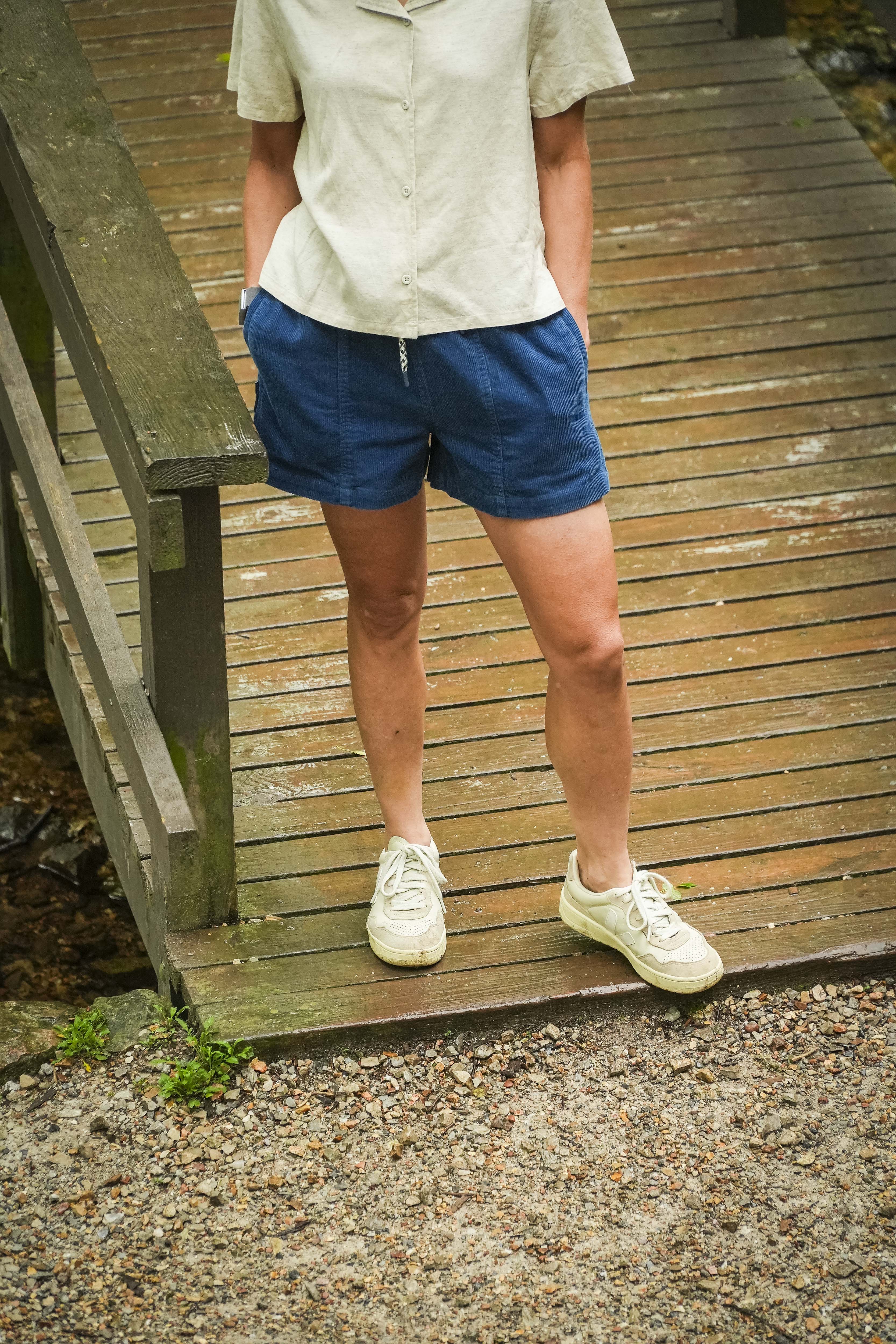 A person wearing BEARDEDGOAT Womens Cord Shorts and a light short-sleeve shirt stands on a wooden bridge with hands in pockets, white sneakers, gravel, and greenery nearby. Their head is not visible in the image.