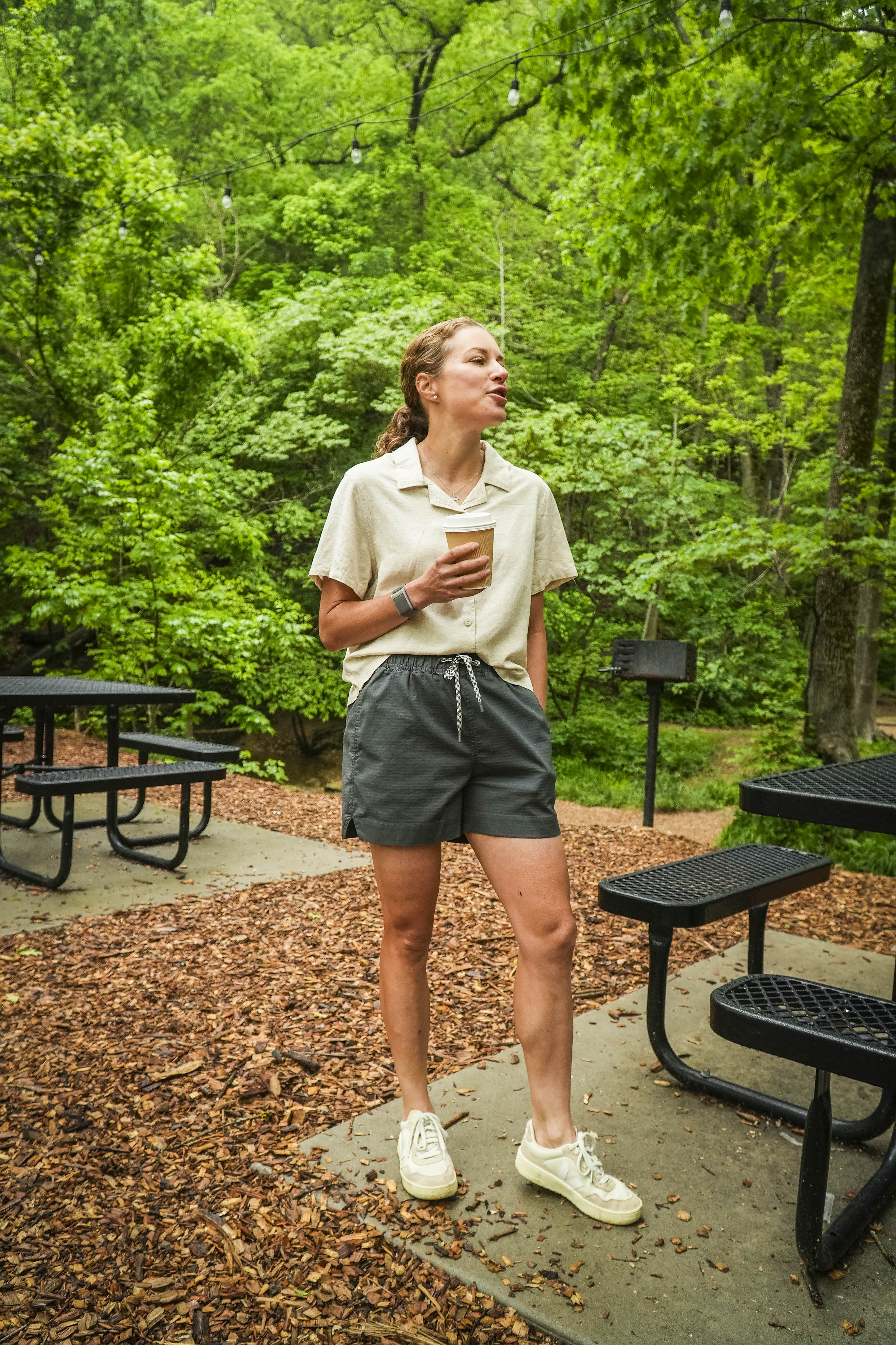 A woman stands at a picnic area surrounded by greenery, holding a coffee cup and wearing BEARDEDGOATs Womens Canopy Short in gray stretch cotton ripstop, paired with an athletic fit light shirt and sneakers, looking relaxedly to the side.