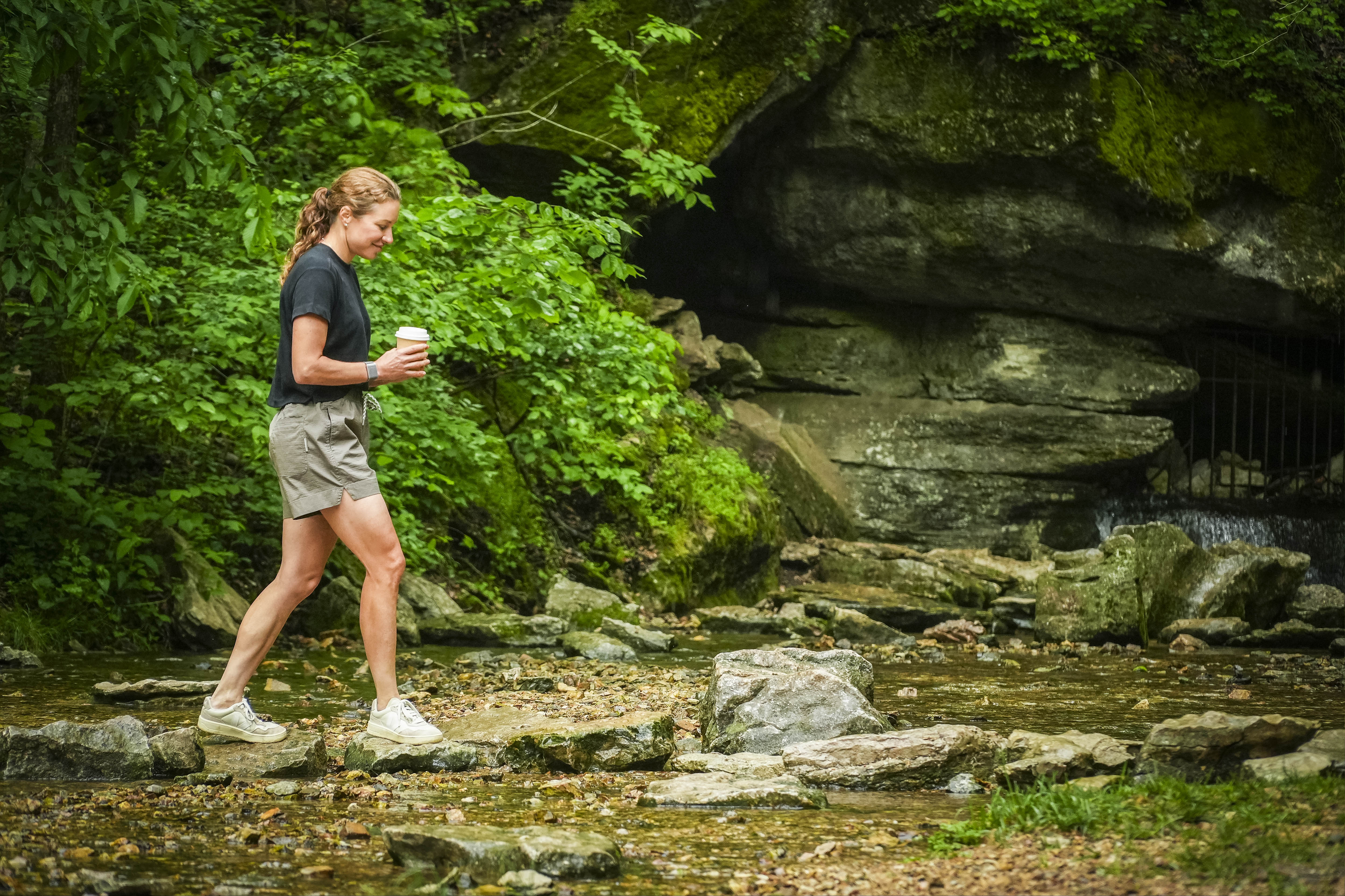 A red-haired woman in a black shirt and BEARDEDGOAT Womens Canopy Short made of stretch cotton ripstop walks across rocks in a shallow creek, holding a cup, with lush green trees and a rocky cave opening behind her.