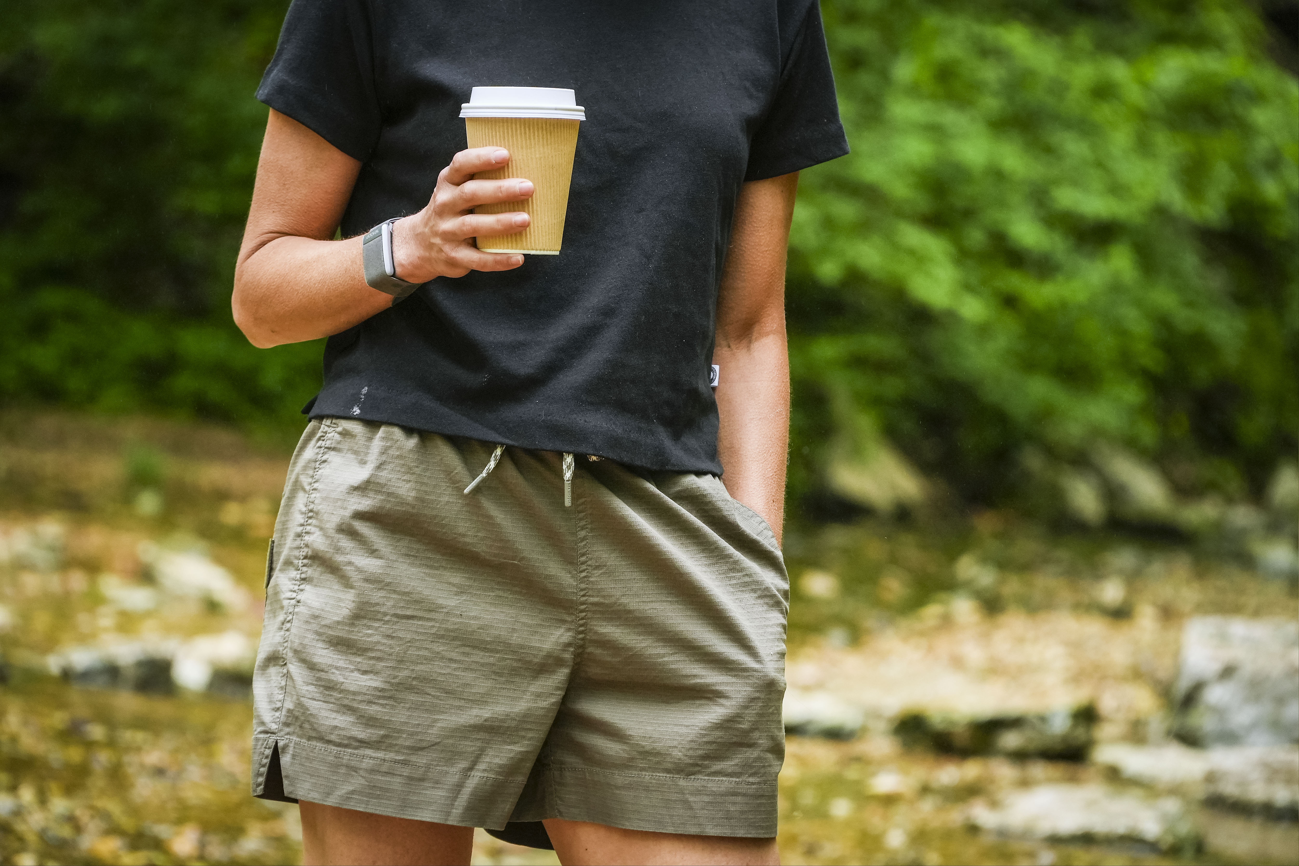 A person wearing BEARDEDGOAT Womens Canopy Short and a black t-shirt stands outdoors with a coffee cup in one hand and the other hand in their pocket, surrounded by green foliage and rocks.