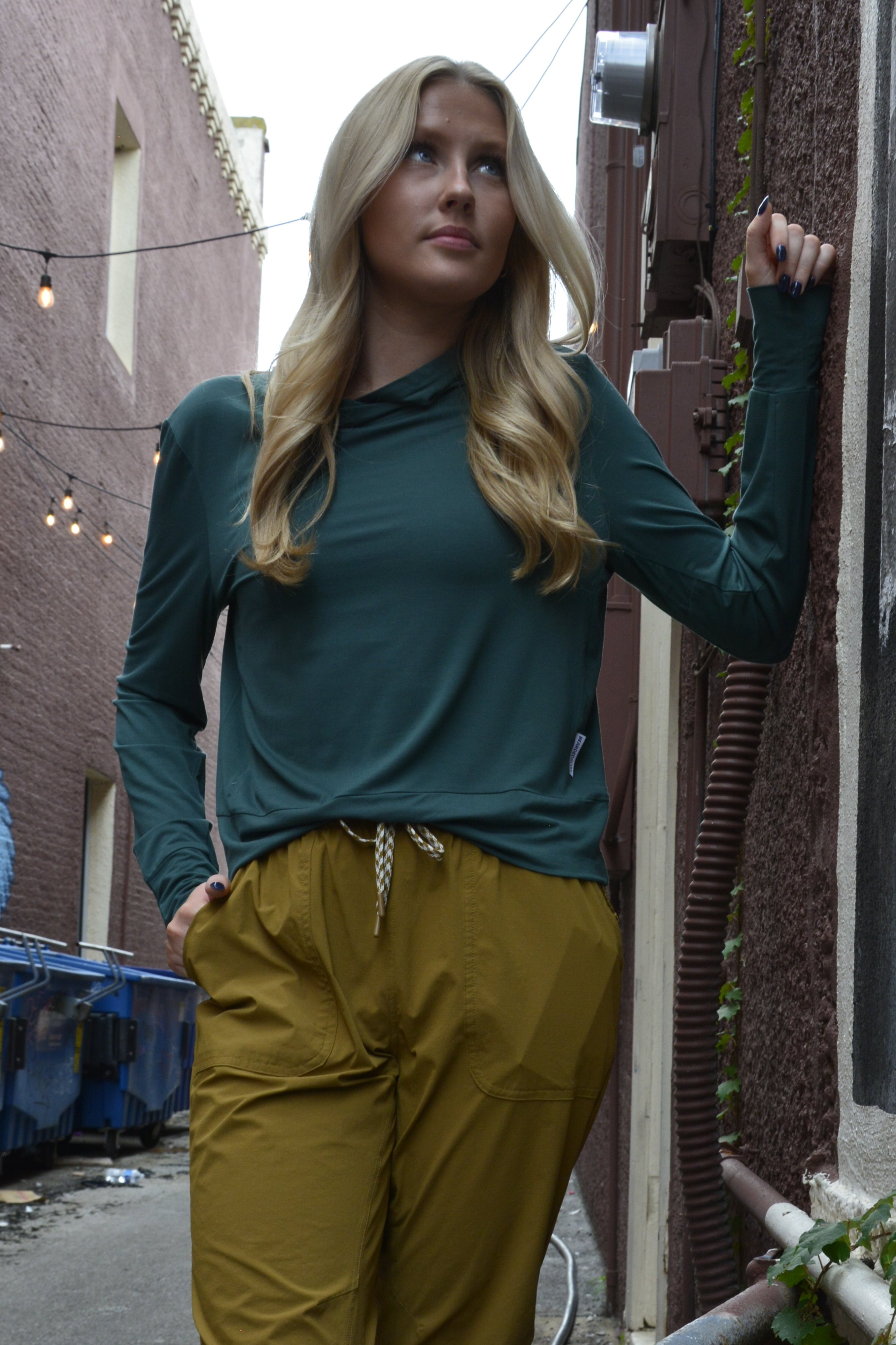 A woman with long blonde hair stands in an alleyway, wearing the BEARDEDGOAT Women’s EverZen™ Bamboo Hoodie in teal and mustard yellow pants. One hand is in her pocket, the other on a wall, with string lights and blue bins around her.