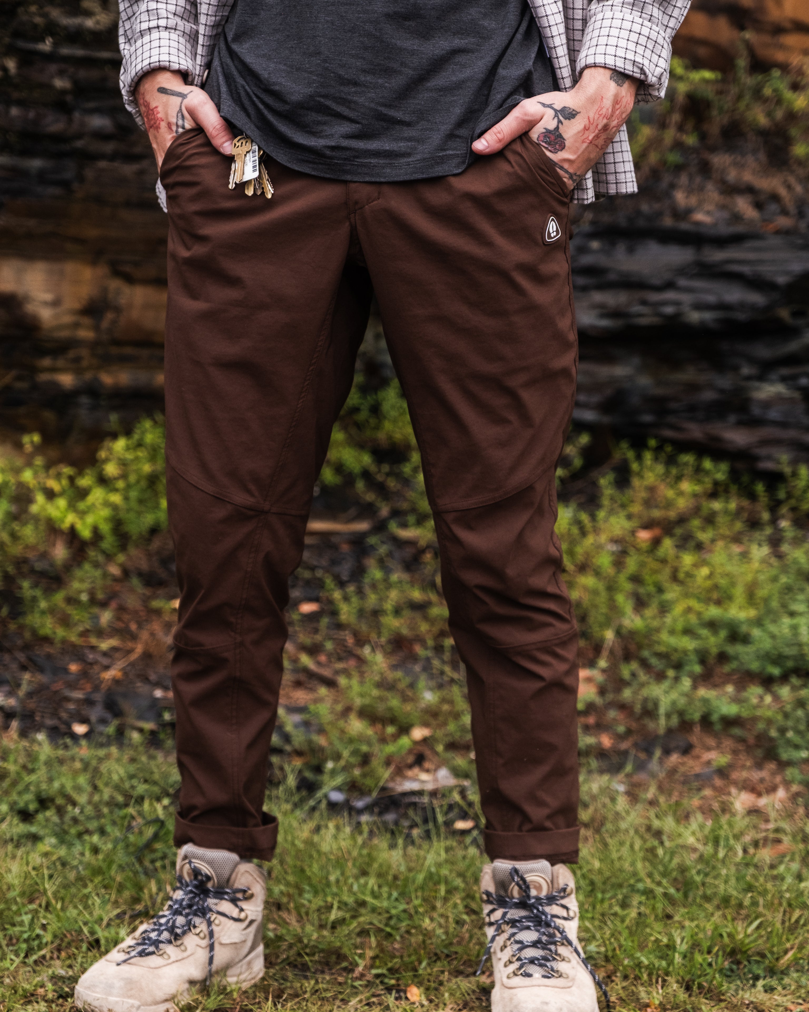 Standing outdoors on grass, hands in pockets, a person wears BEARDEDGOAT NEW—ROVER™ Pant, a dark shirt, plaid overshirt, light hiking boots, and holds keys. Only the lower half of the body is visible.