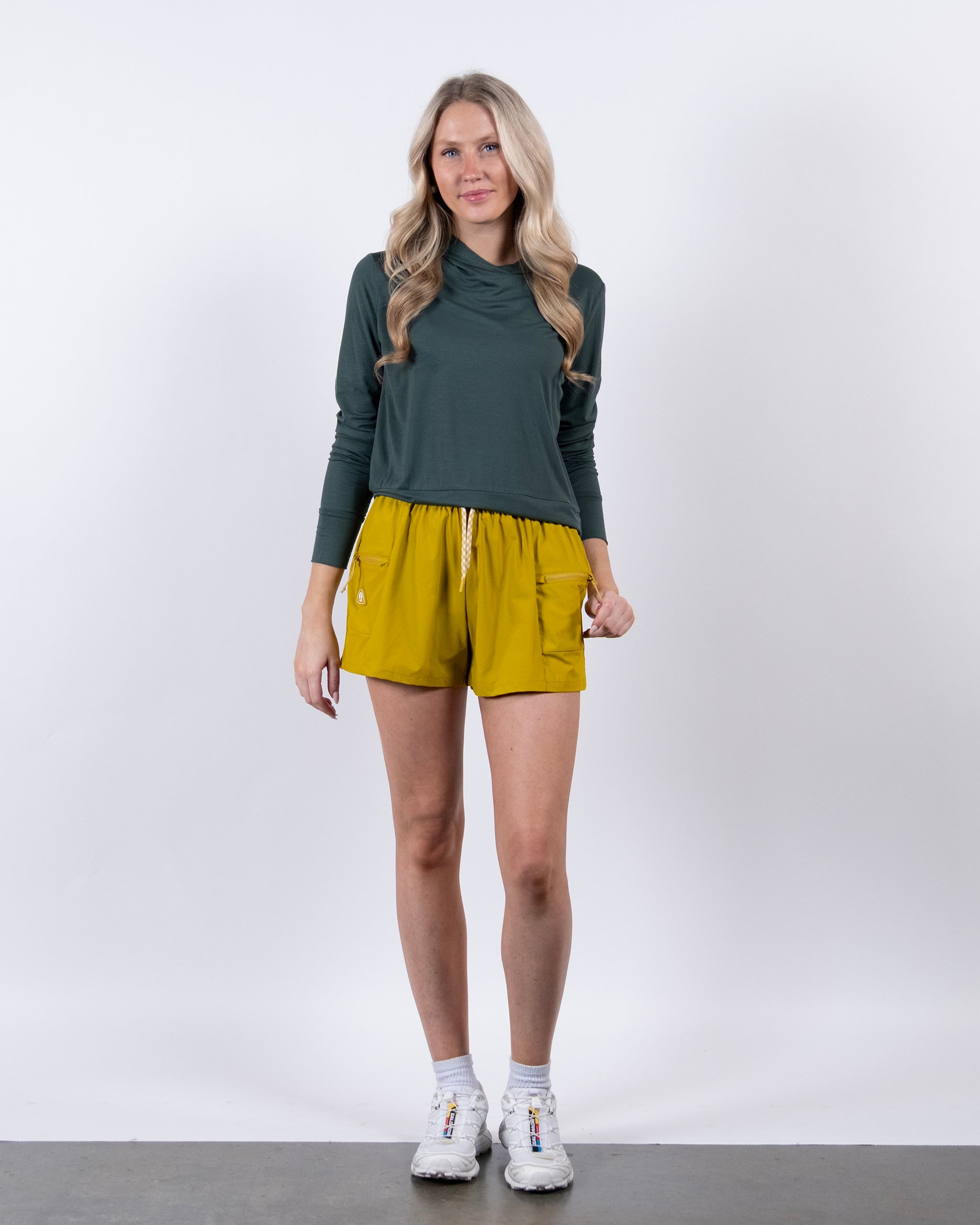 A woman with long blonde hair stands against a white background wearing the BEARDEDGOAT Women’s EverZen™ Bamboo Hoodie in dark green, paired with yellow drawstring shorts and white sneakers, facing forward with a relaxed expression.