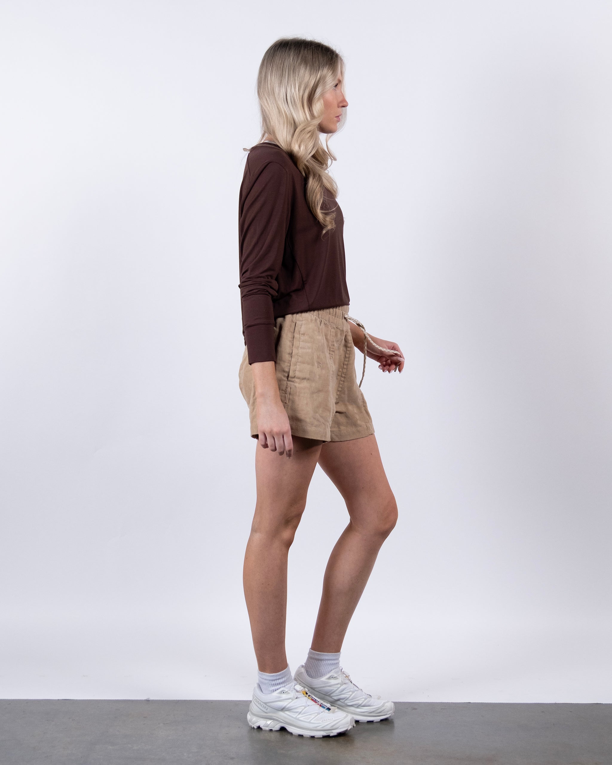 A woman with long blonde hair stands in profile against a plain white background, wearing the BEARDEDGOAT Women’s EverZen™ Bamboo Long Sleeve in brown, tan shorts, white socks, and chunky white sneakers.