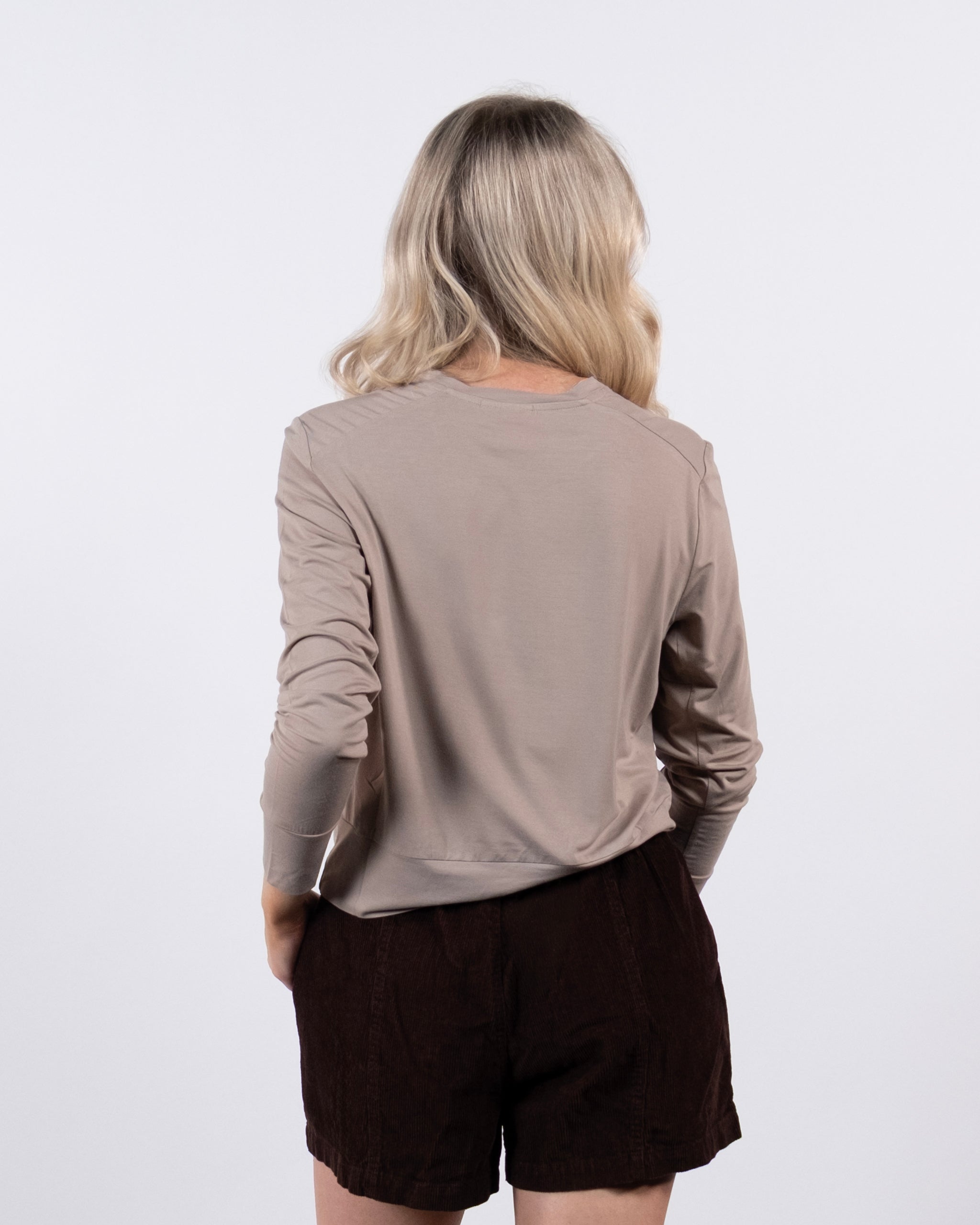 A person with long, wavy blonde hair stands facing away from the camera, wearing a BEARDEDGOAT Women’s EverZen™ Bamboo Long Sleeve in beige and dark brown shorts against a plain white background.