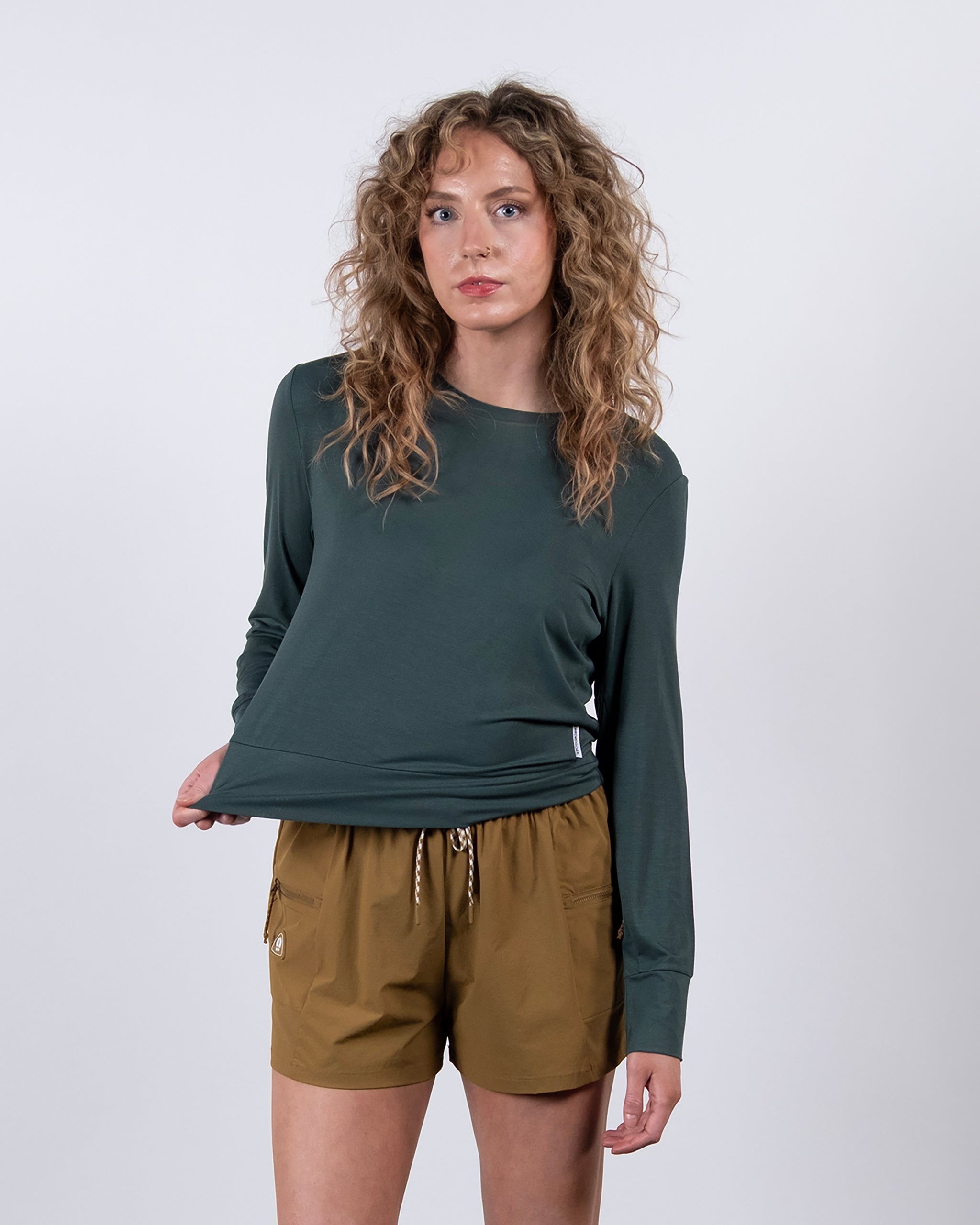 A woman with curly light brown hair wears the BEARDEDGOAT Women’s EverZen™ Bamboo Long Sleeve in dark green and tan drawstring shorts, standing against a plain white background. She holds her shirt’s side and looks at the camera.