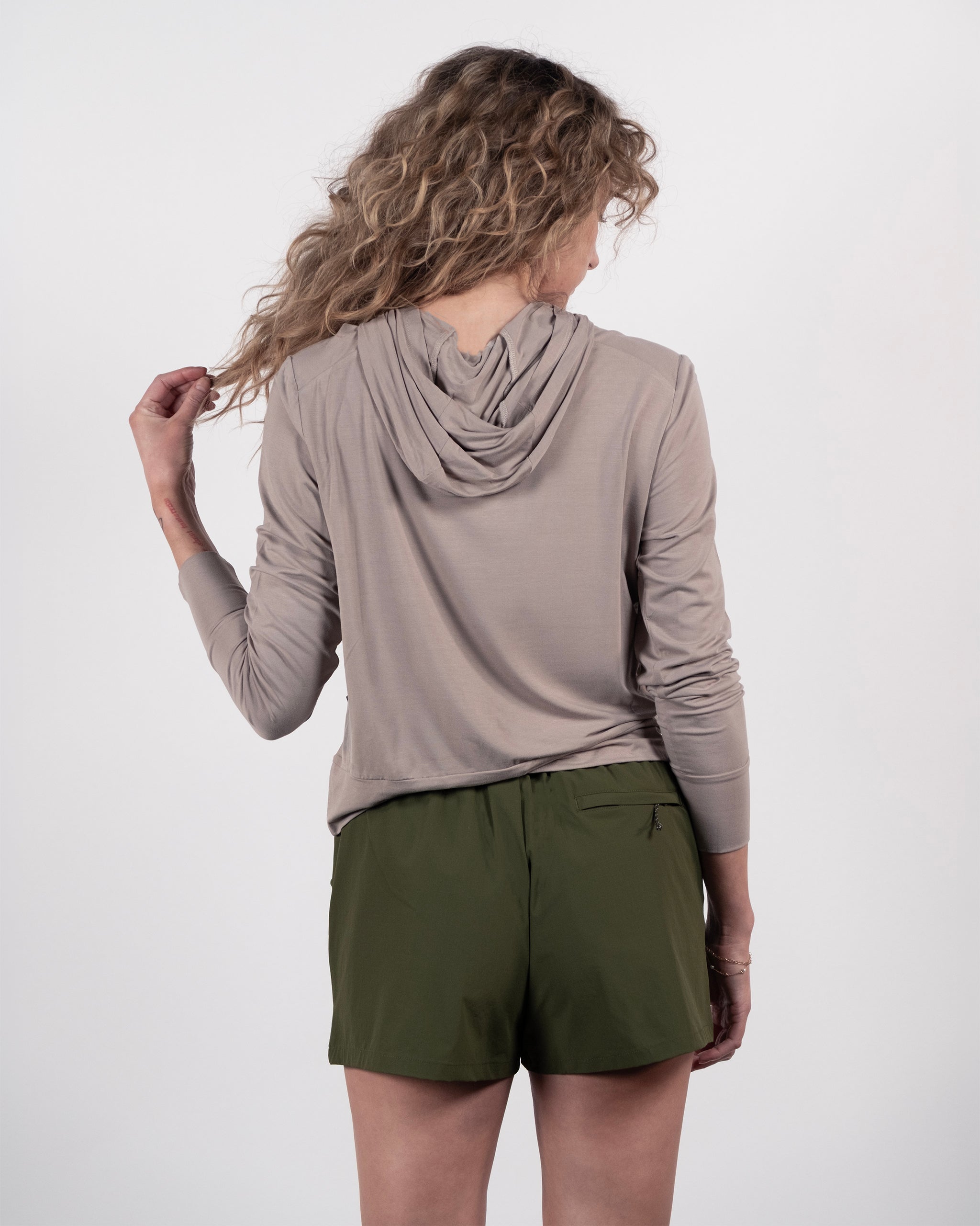A person with wavy hair, facing away from the camera and gently holding a strand of hair, wears the BEARDEDGOAT Women’s EverZen™ Bamboo Hoodie in light taupe paired with olive green shorts.