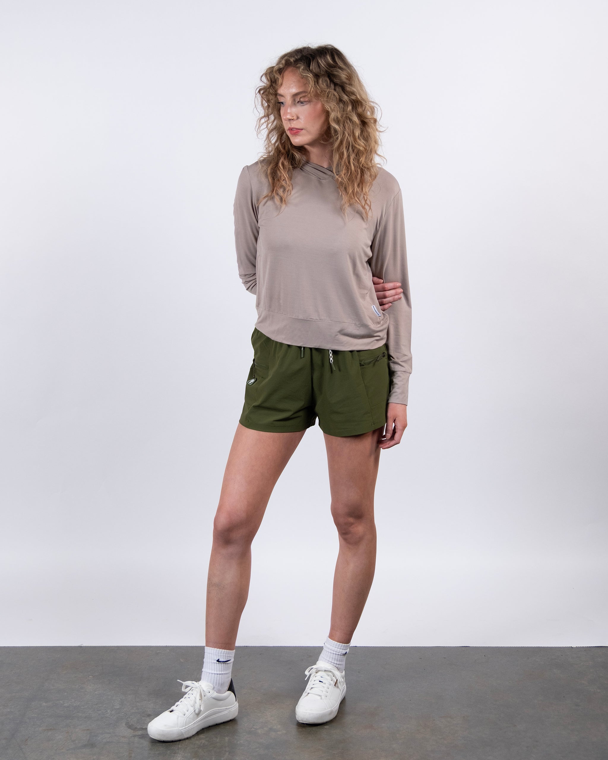 A person with curly hair wears a light beige BEARDEDGOAT Women’s EverZen™ Bamboo Hoodie, olive green shorts, white socks, and white sneakers, standing on a gray floor against a plain white background.