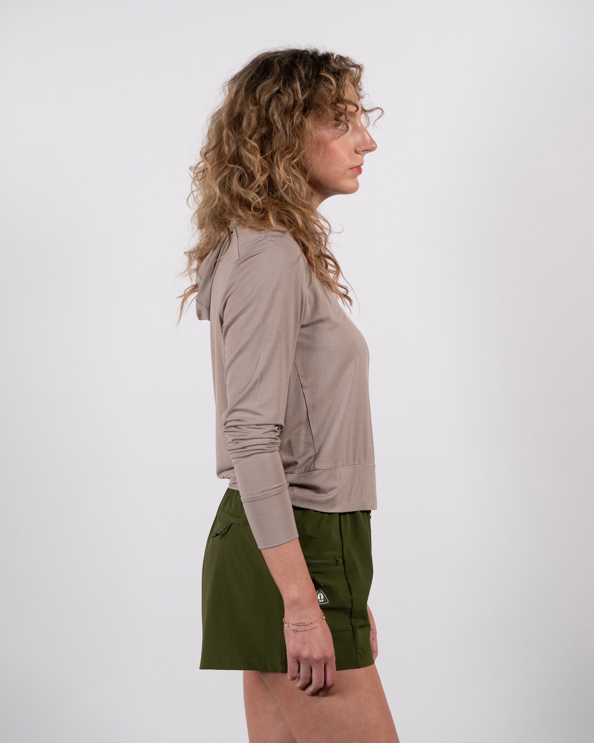 A woman with curly hair wears the eco-conscious BEARDEDGOAT Women’s EverZen™ Bamboo Hoodie in taupe and olive green shorts, standing in profile against a plain white background.