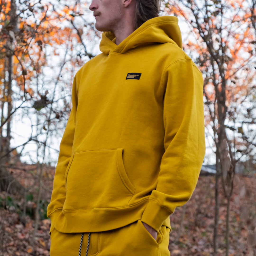 A man wearing the beardedgoat Men's Void Hoodie in Golden Palm stands outdoors among autumn leaves. The heavyweight cotton fleece keeps him warm as he looks to the side, hands tucked in his pockets.