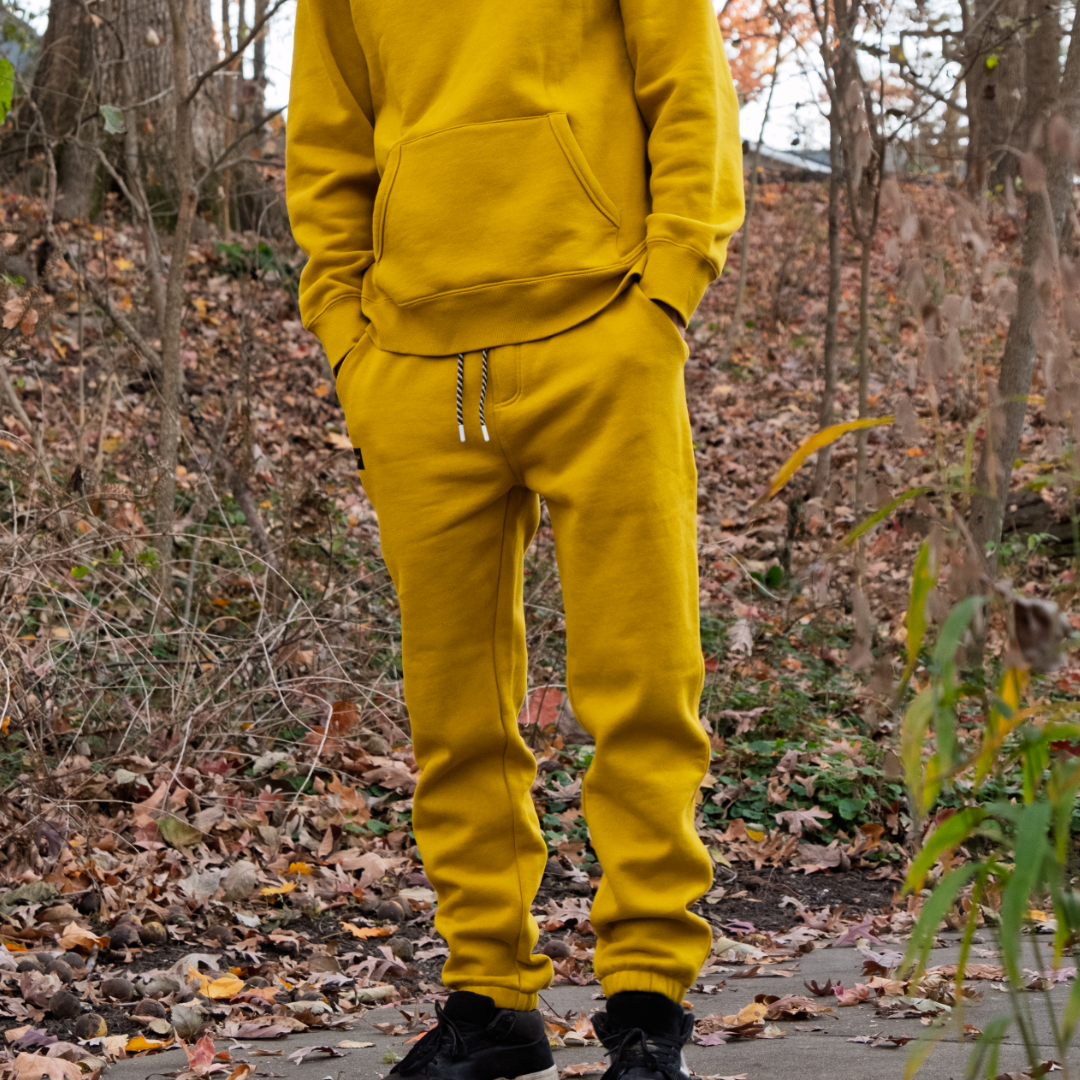 In beardedgoat’s Men’s Void Sweatpants and a matching yellow hoodie, a person stands on a leafy path amid autumn trees, with only their body visible and face unseen.