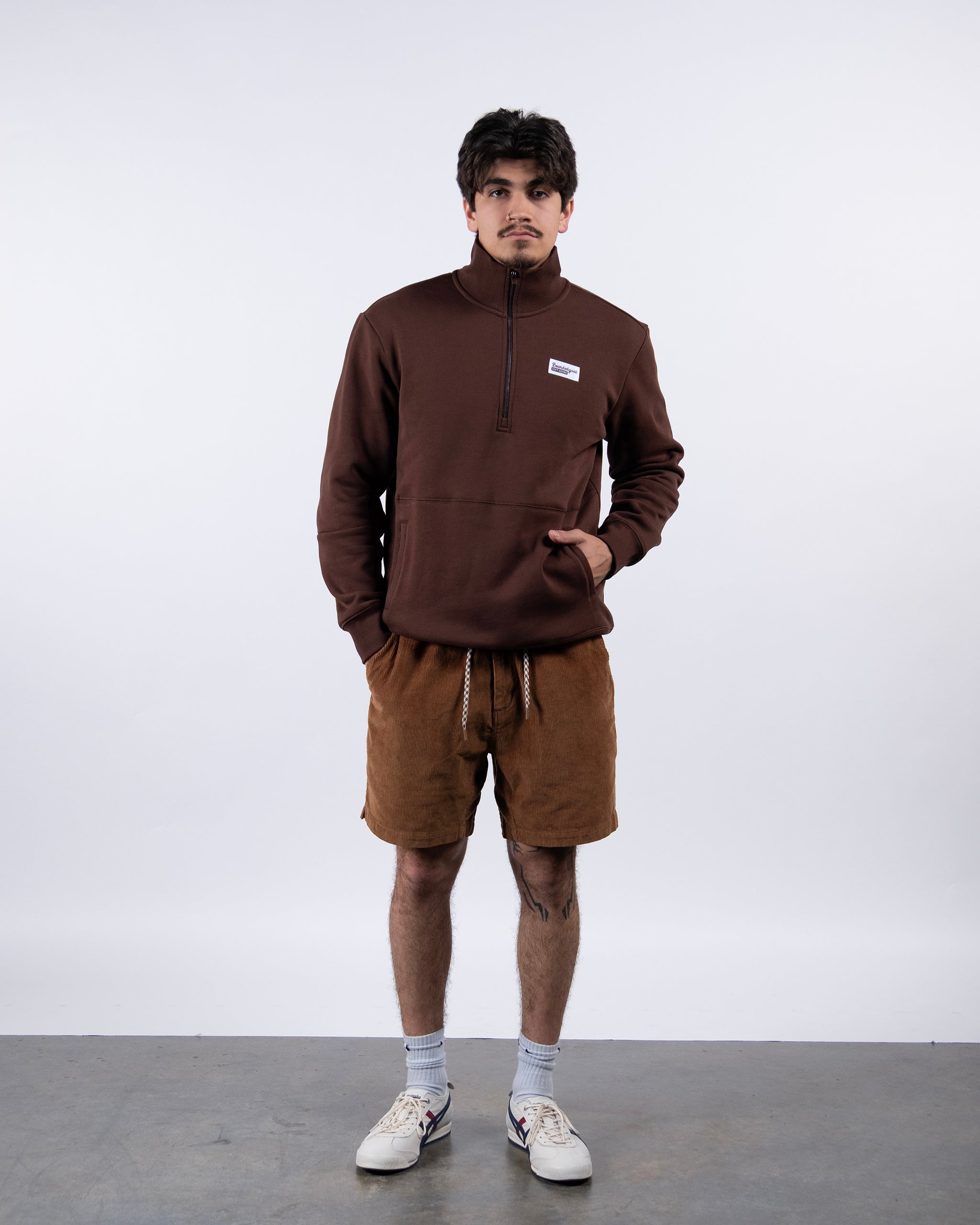 A man stands against a plain white background wearing the BEARDEDGOAT NEW — VOID™ Half Zip, brown drawstring shorts, white socks, and white sneakers. His hands are in his pockets and he looks directly at the camera.
