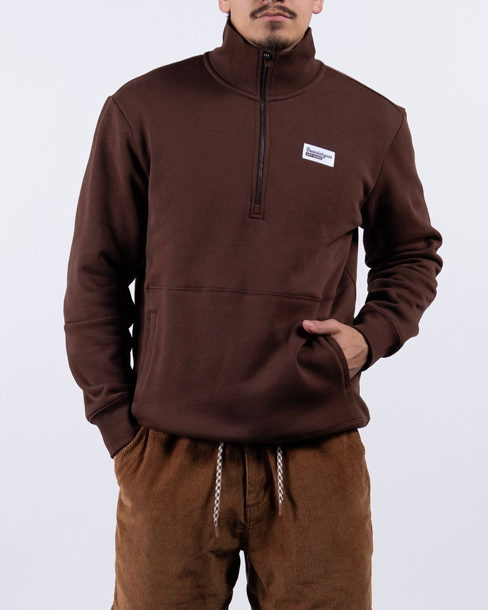 A man stands against a plain white background wearing the BEARDEDGOAT NEW — VOID™ Half Zip, brown drawstring shorts, white socks, and white sneakers. His hands are in his pockets and he looks directly at the camera.